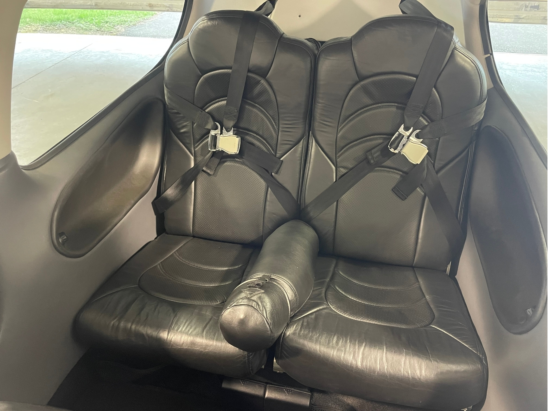 Two black leather airplane seats with shoulder and lap belts fastened window visible to the side in a small aircraft cabin