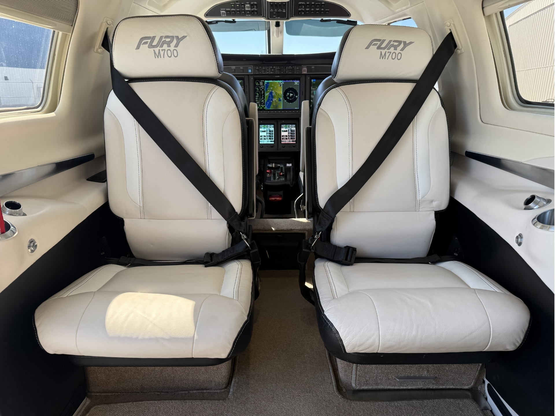 Front cabin view of two beige captain seats in a small aircraft with seat belts fastened and a glass cockpit dashboard ahead