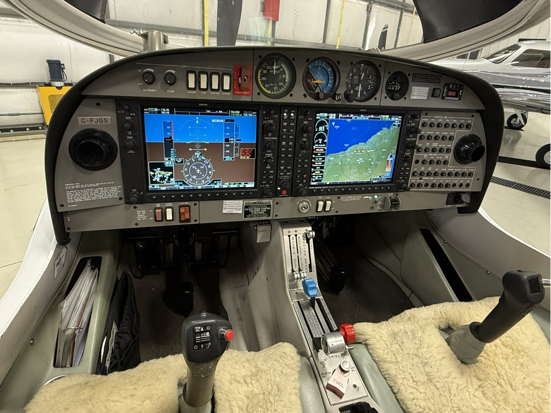 Cockpit of a small airplane with two large glass displays, analogue gauges, and a cluttered center console in a hangar bay.