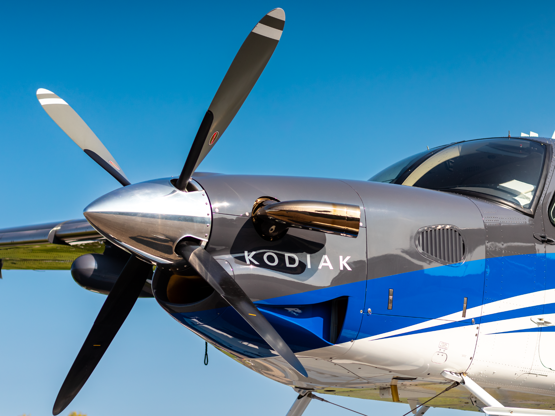 Close up of a propeller driven aircraft with a glossy gray nose two blade propeller and cockpit windows against a blue sky