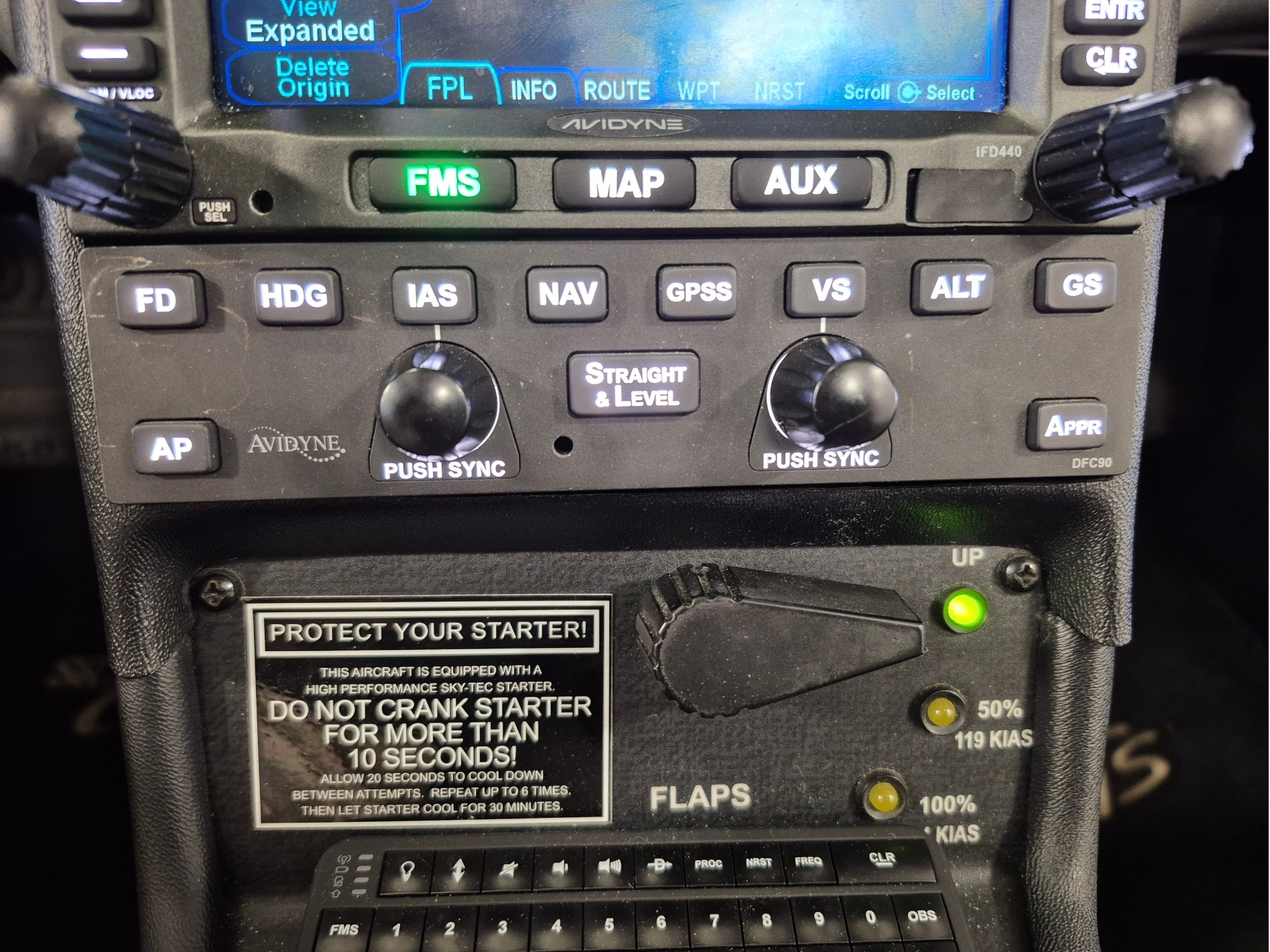Cockpit panel with a blue GPSnavigation screen above and labeled control buttons FMS green MAP AUX row of small square buttons FD HDG IAS NAV GPS VS ALT GS two large knobs and a Straight Level button in the center warning plaque about starter below
