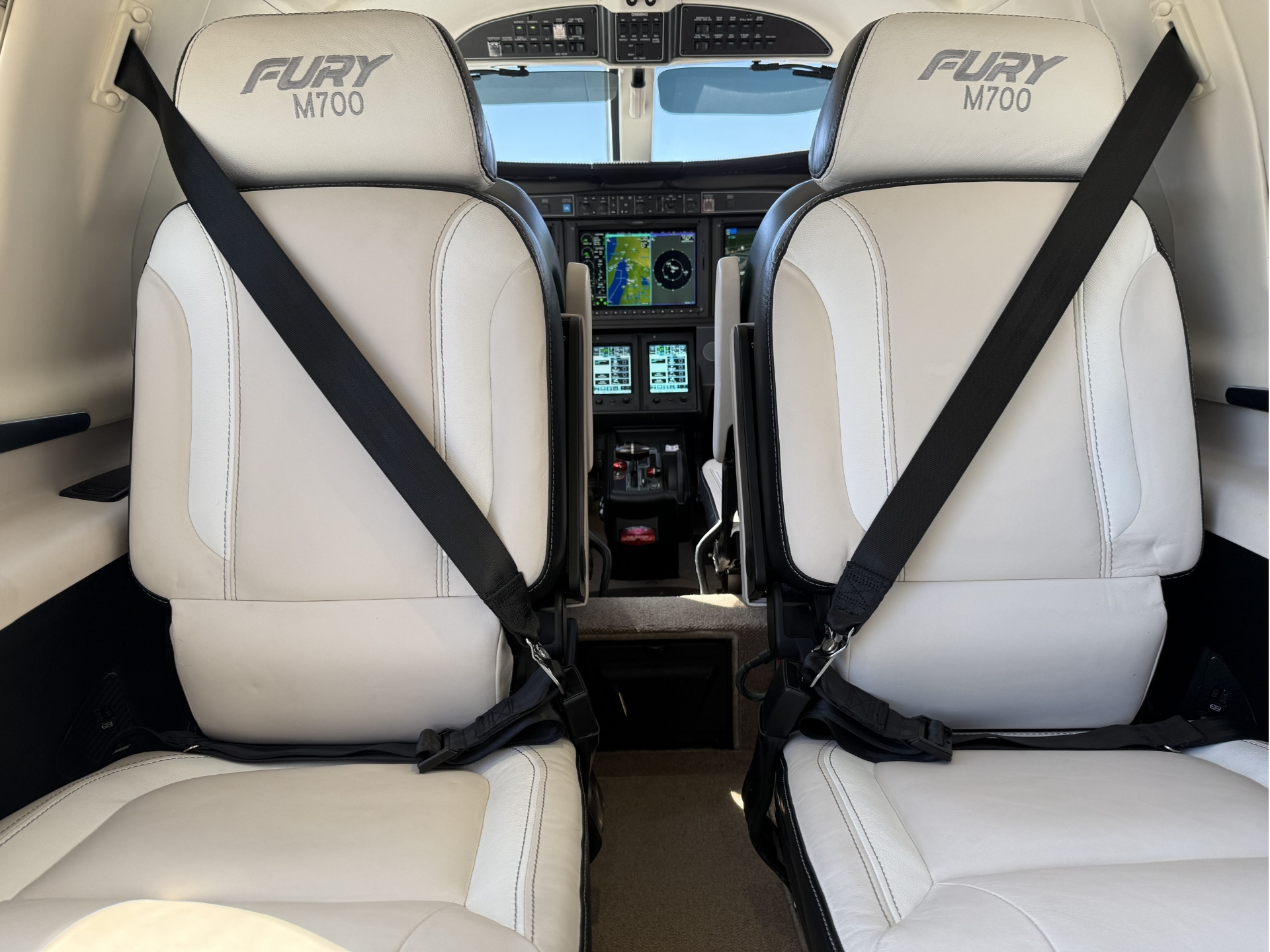 Front cockpit of a small airplane viewed from behind two cream captain seats with buckled black seat belts and a digital cockpit display in the center