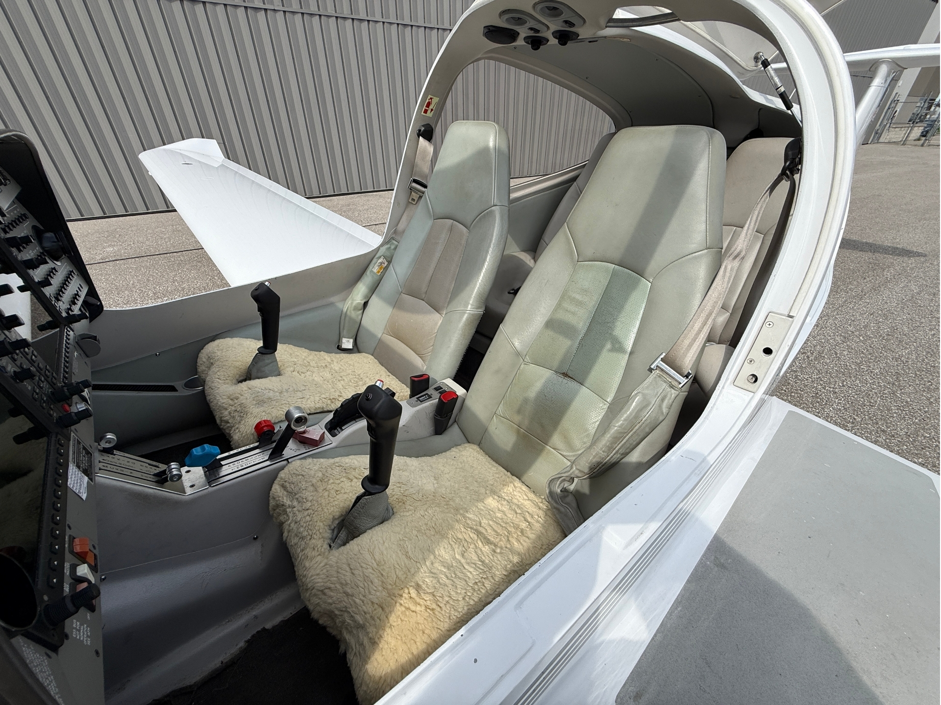 Interior view of a small airplane cockpit with two beige seats, control yokes, and throttle levers; fluffy cream-colored carpet on the floor; open door showing the tarmac outside a hangar.