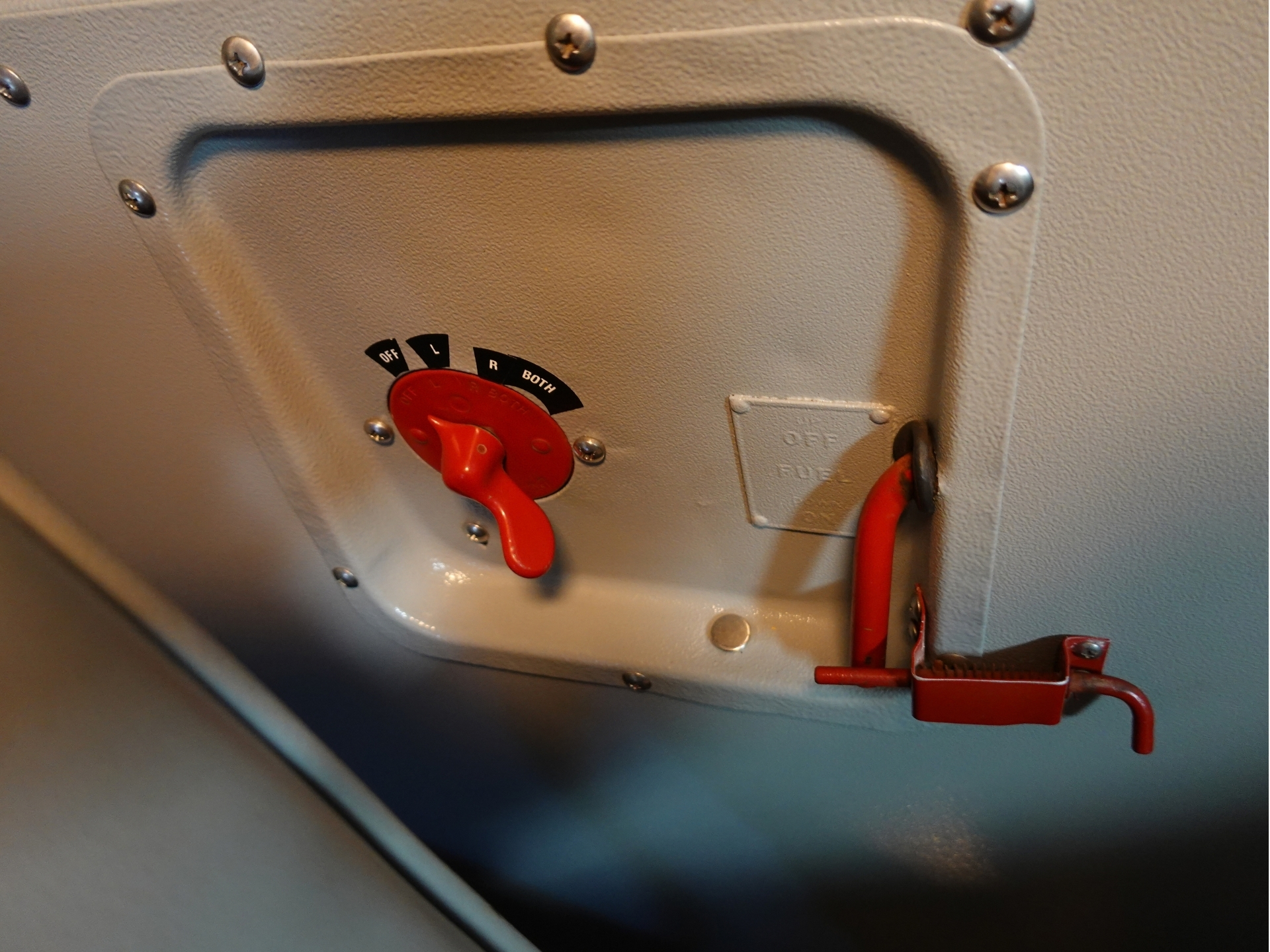 Close up of a red toggle lever labeled OFF L R BOTH on a gray metal cockpit panel with screws nearby