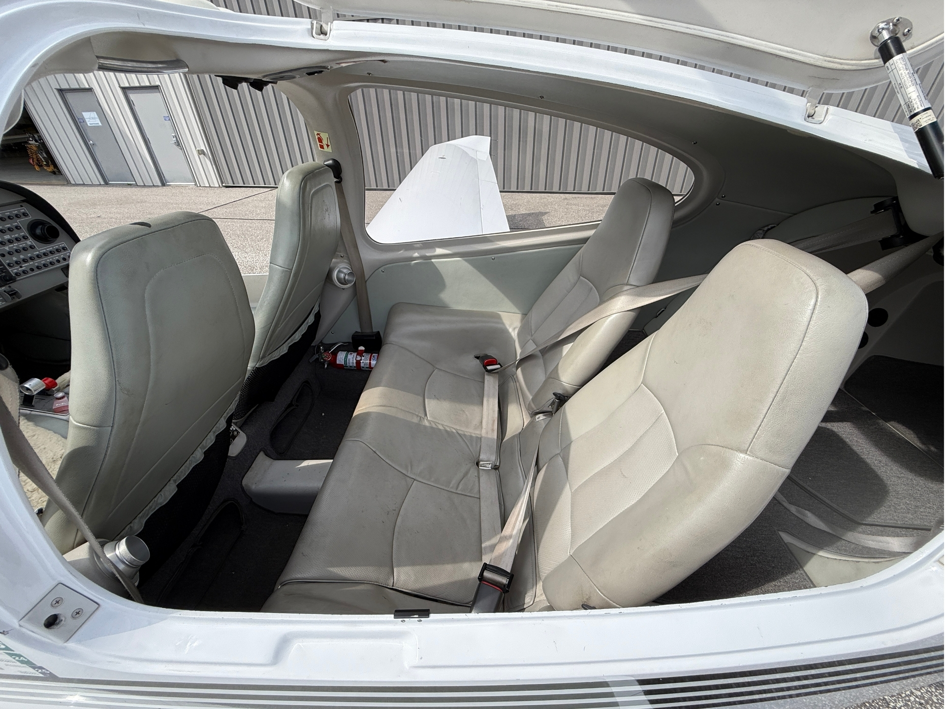 Interior of a small light aircraft showing beige seats, cockpit controls on the left, and an open canopy view of the hangar outside.