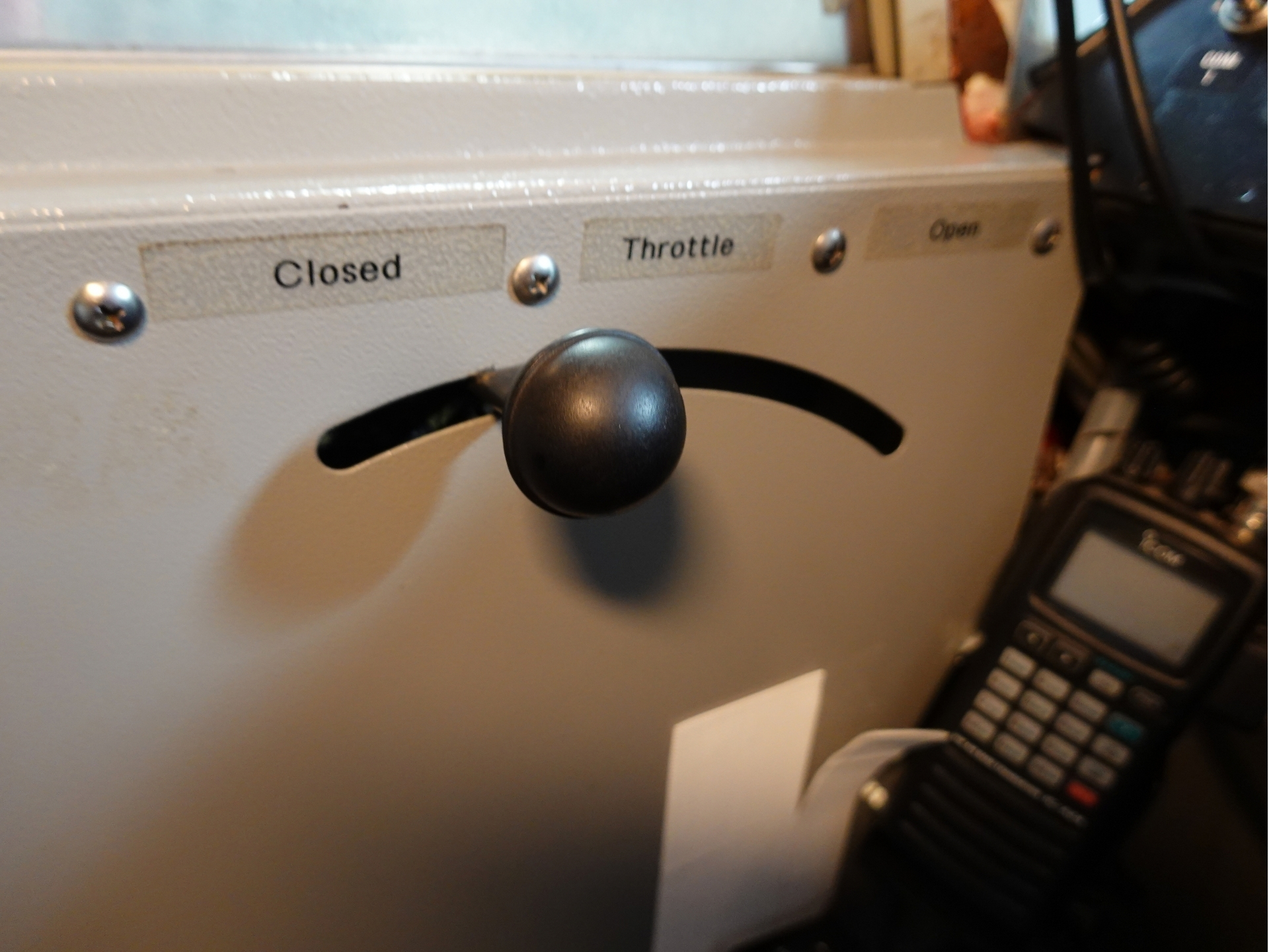 Close up of a gray control panel with a black lever knob and three labels Closed Throttle Open on a machine or cockpit panel