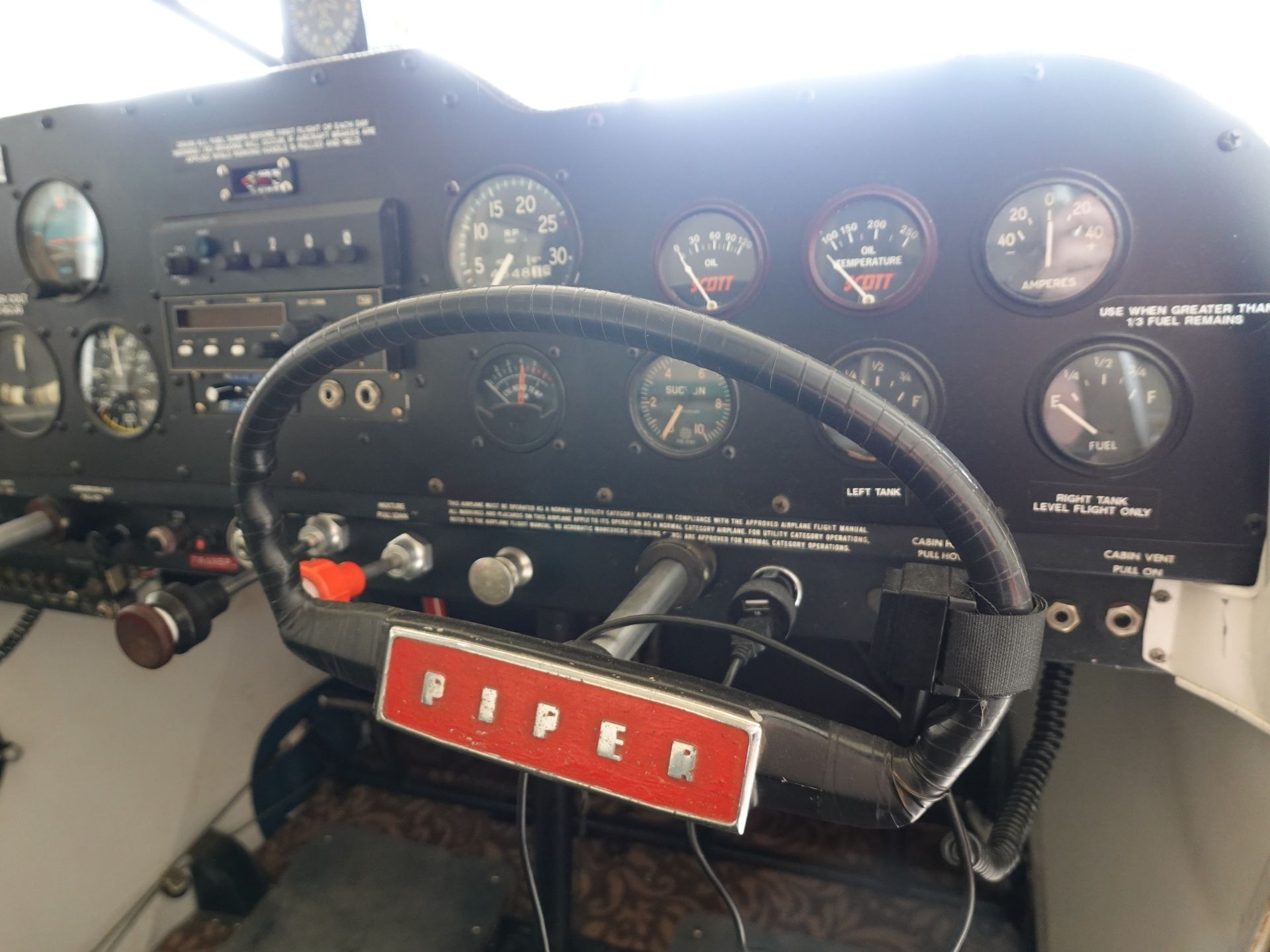 Close-up of a small aircraft cockpit with a yoke in the foreground and round flight instruments on the panel, including fuel and oil gauges, labeled Piper on the control area.