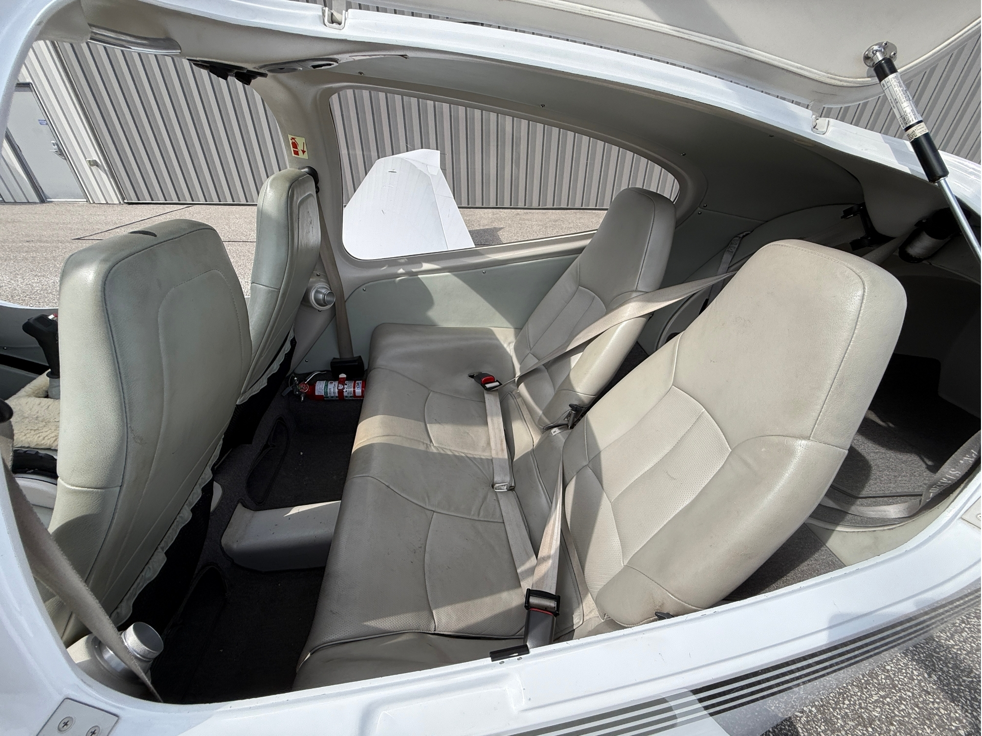Interior of a small light aircraft showing two rows of beige seats with seat belts and an open canopy above.