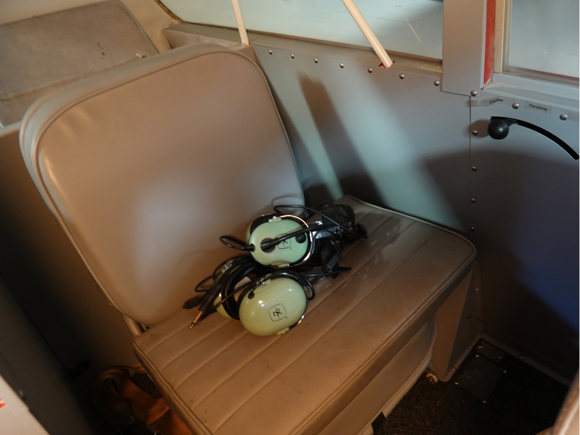 A pair of green aviation headsets with cords on a beige airplane seat beside a cockpit window panel