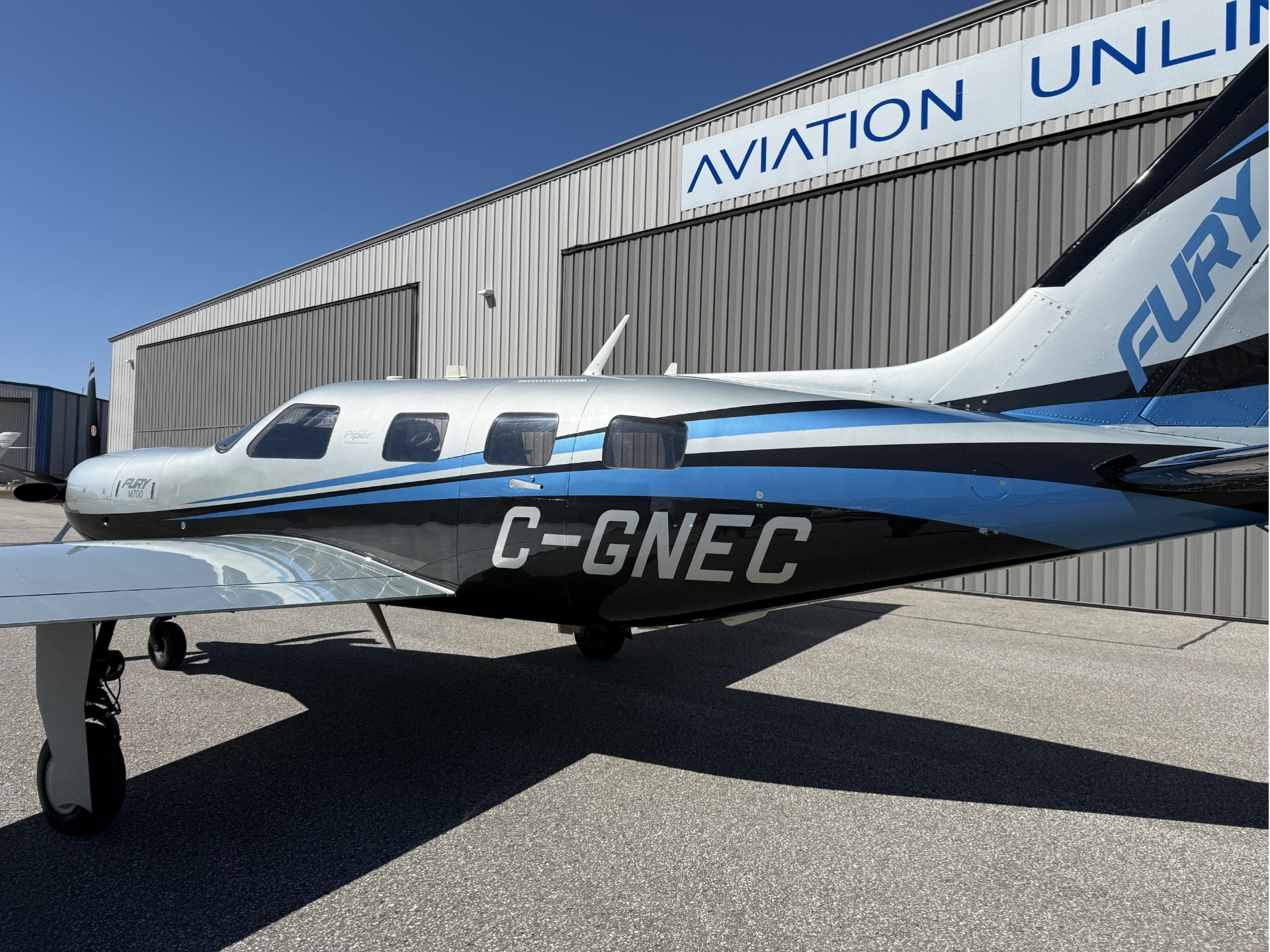 Private turboprop aircraft with light gray and blue livery parked on tarmac beside a corrugated hangar with an Aviation Unlimited sign