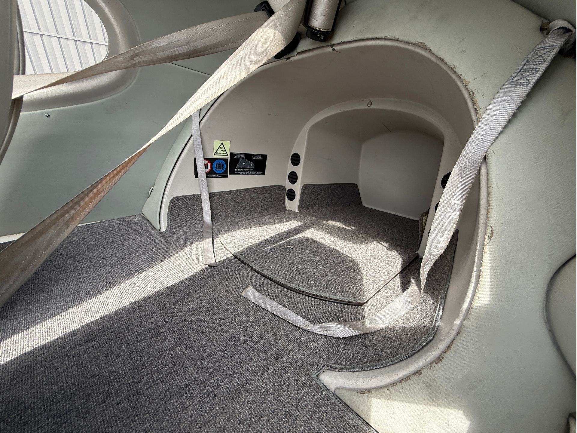 Inside a small aircraft cabin showing gray carpeting, seat belts draped across a curved wall and floor, with scuffed interior paneling.