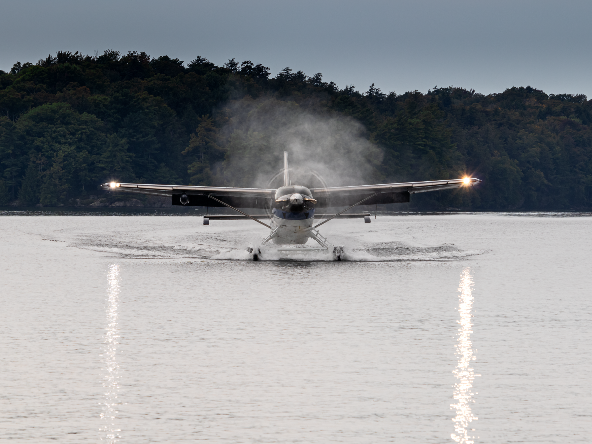 Front view of a seaplane landing on a lake with water spray and a forested shoreline behind it