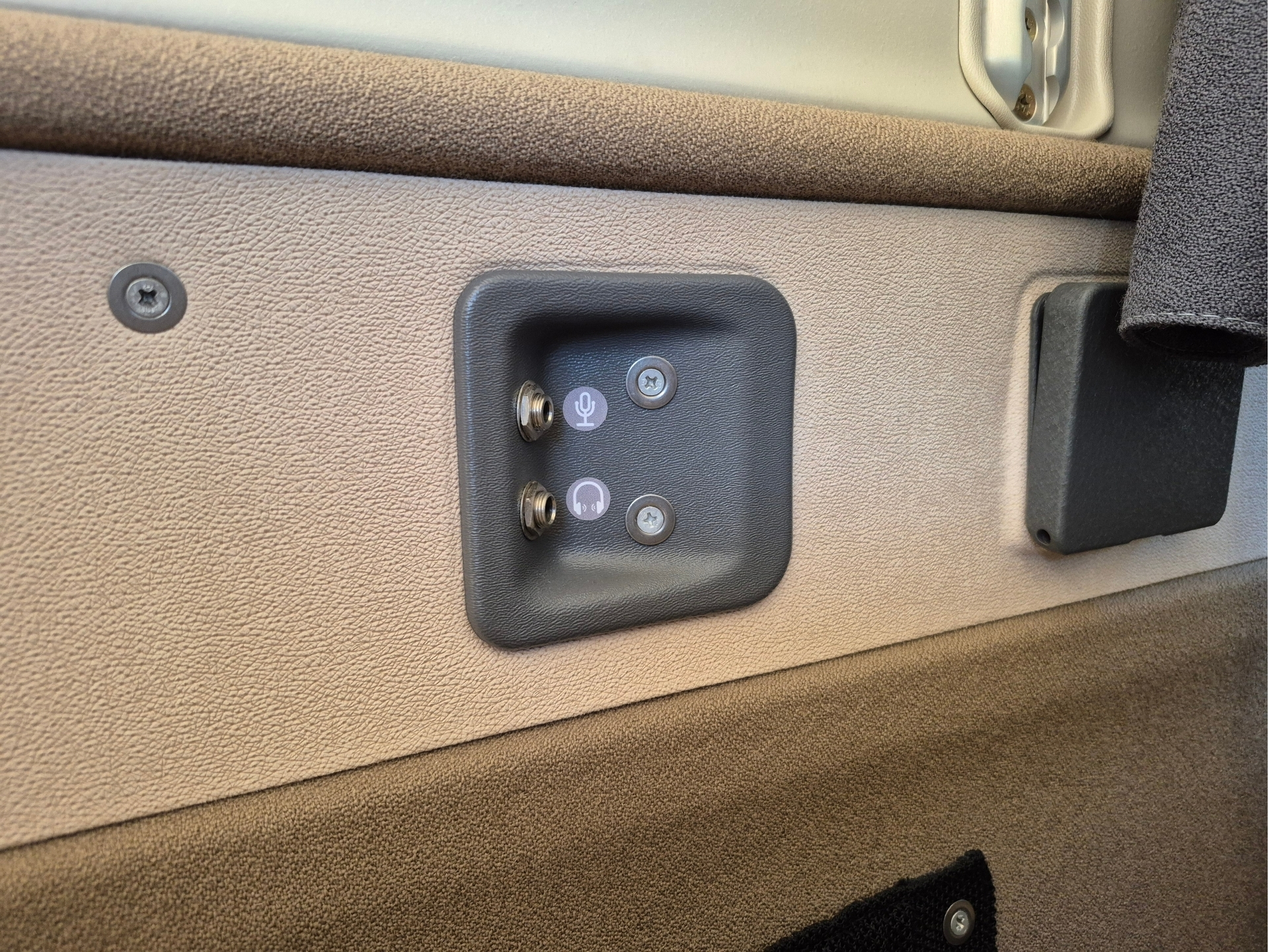Close up of a beige car interior panel showing a black control plate with a microphone and headset jack ports on a textured surface