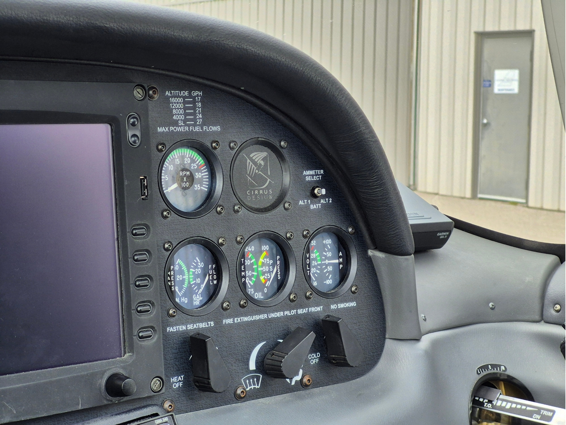 Close up of a small aircraft cockpit panel with multiple gauges including an RPM gauge and Cirrus Design emblem with a hangar outside the window