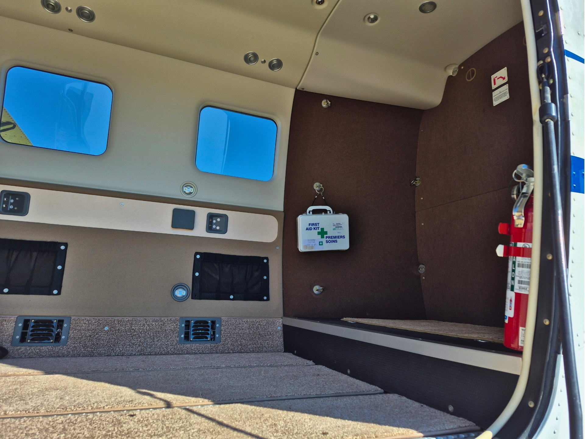Interior of a small aircraft cabin with beige walls a brown side panel a mounted first aid kit and a red fire extinguisher by the door