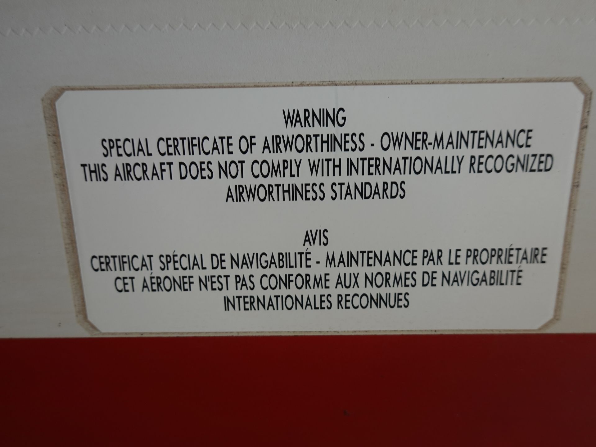 Warning sign about airworthiness standards in multiple languages; large text block on white panel bordered in beige, mounted on a light wall.
