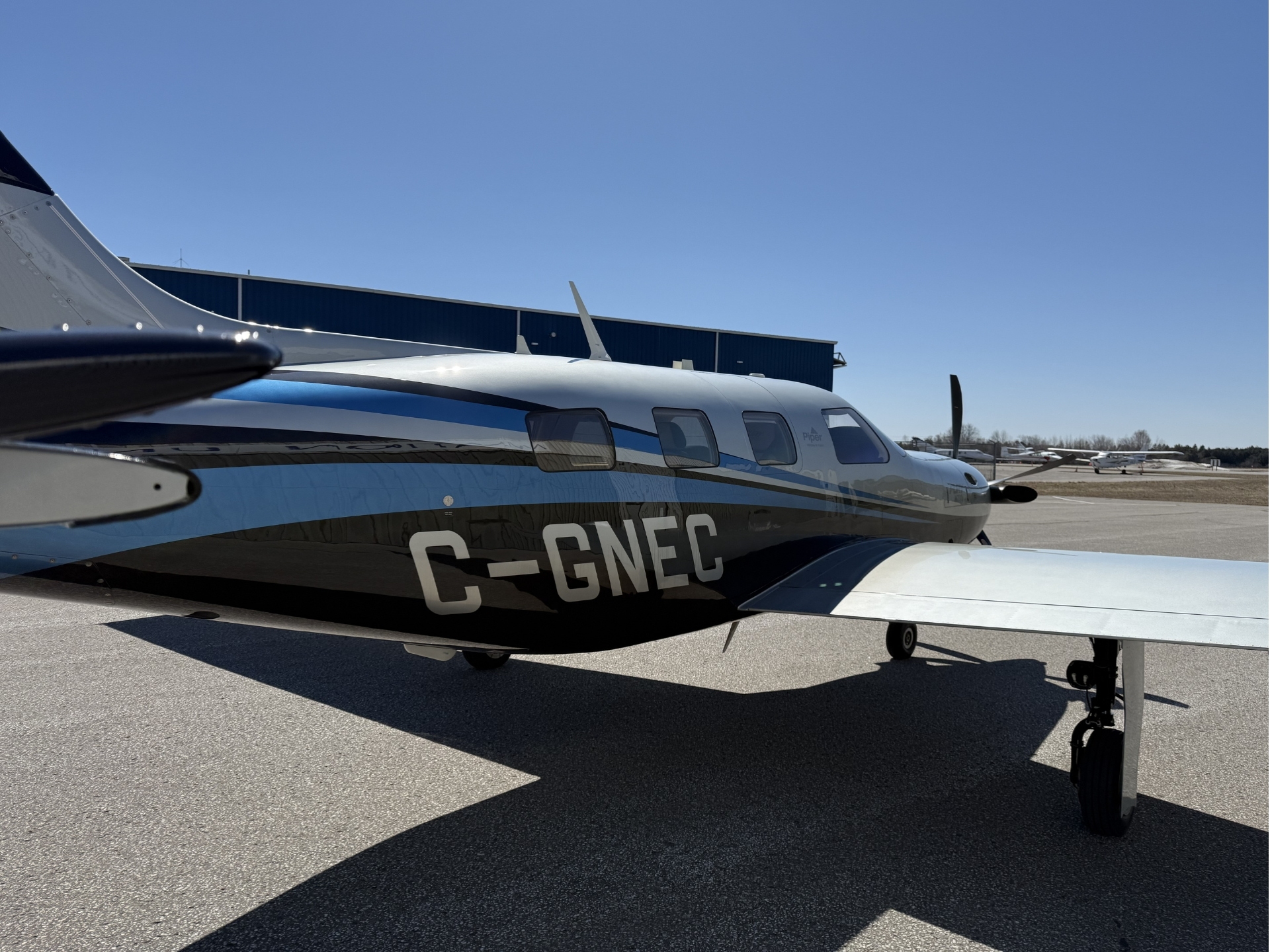Close up of a white and blue private airplane on a tarmac registration C GNEC with blue hangar in the background and clear blue sky above