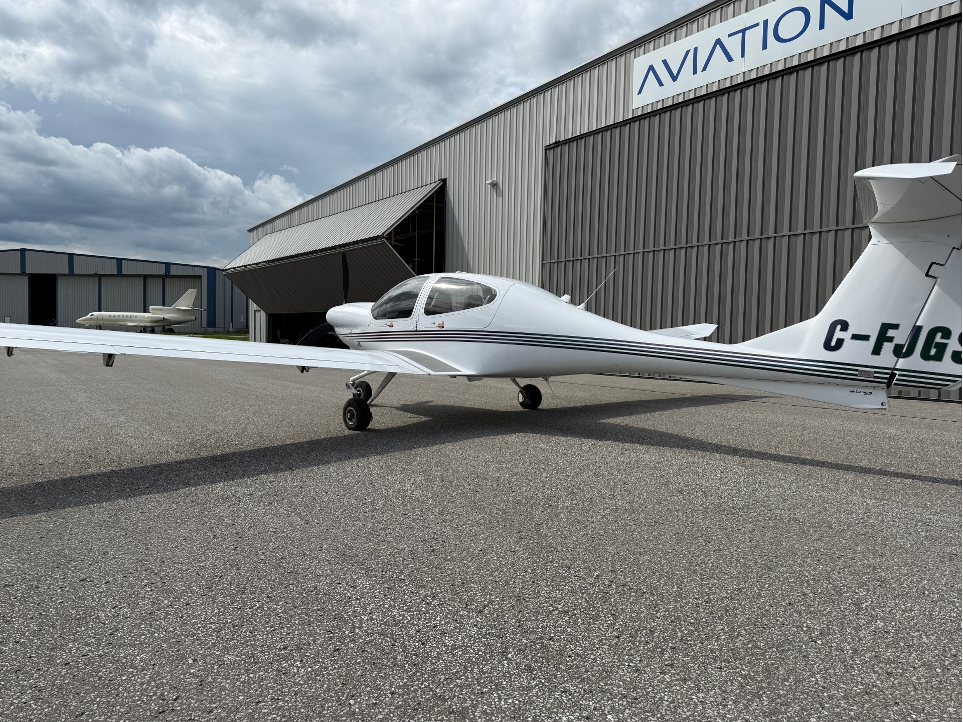 Small white single-engine airplane on the tarmac beside a gray aviation hangar with a large AVIATION sign in the background
