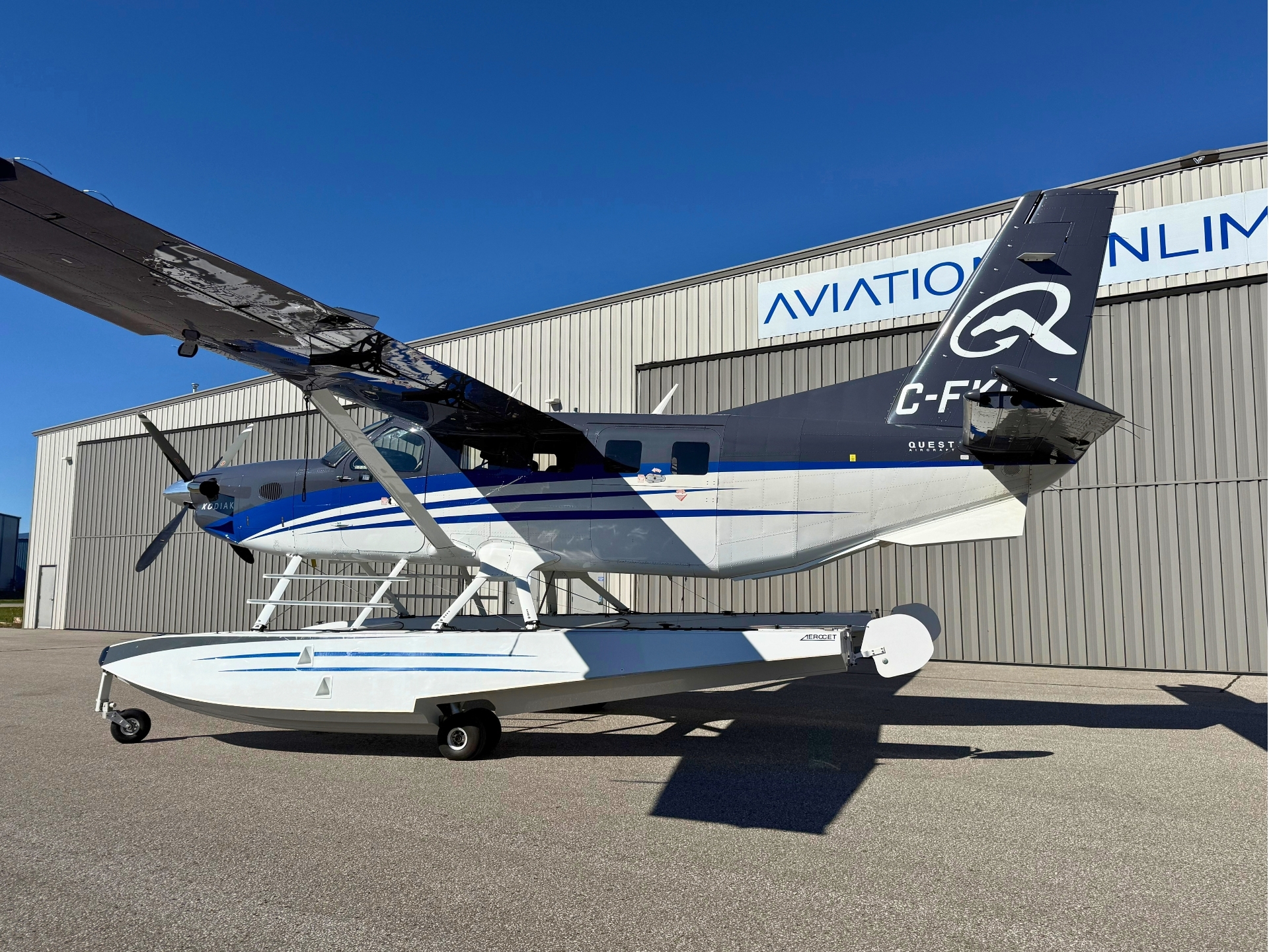 Seaplane on a wheeled trailer next to a hangar blue and white body with bold tail logo and large wing under a clear blue sky