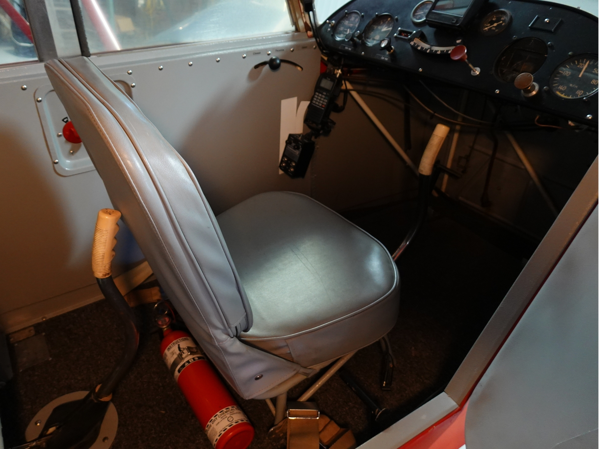Cockpit of a small aircraft showing a gray pilot seat instrument panel on the right and a red fire extinguisher on the floor nearby