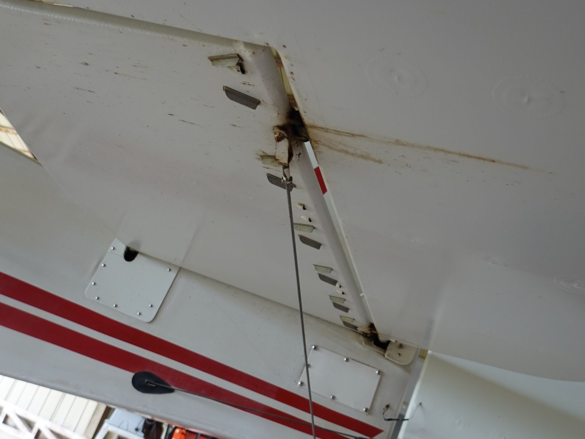 Close-up of a white aircraft wing with rust and exposed fasteners along a panel seam and a tethered cable.