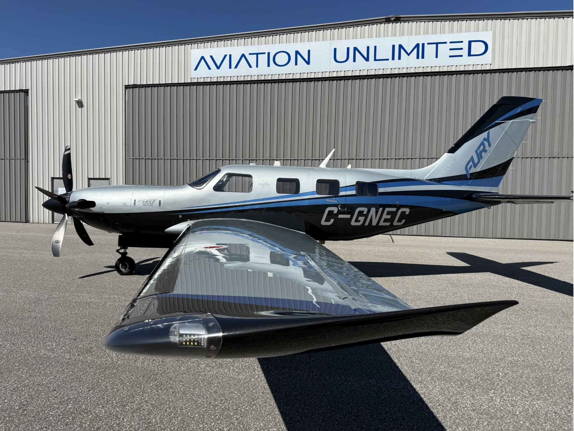 Private silver blue Piper M700 airplane parked in front of a hangar with a white AVIATION UNLIMITED sign above From the cockpit wing perspective