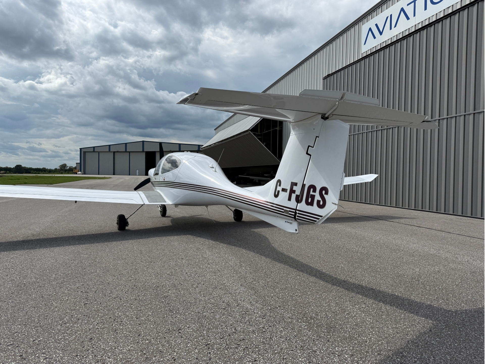 Small white single-engine high-wing airplane on a paved tarmac in front of a gray hangar under a cloudy sky; registration C-FJGS visible on fuselage.