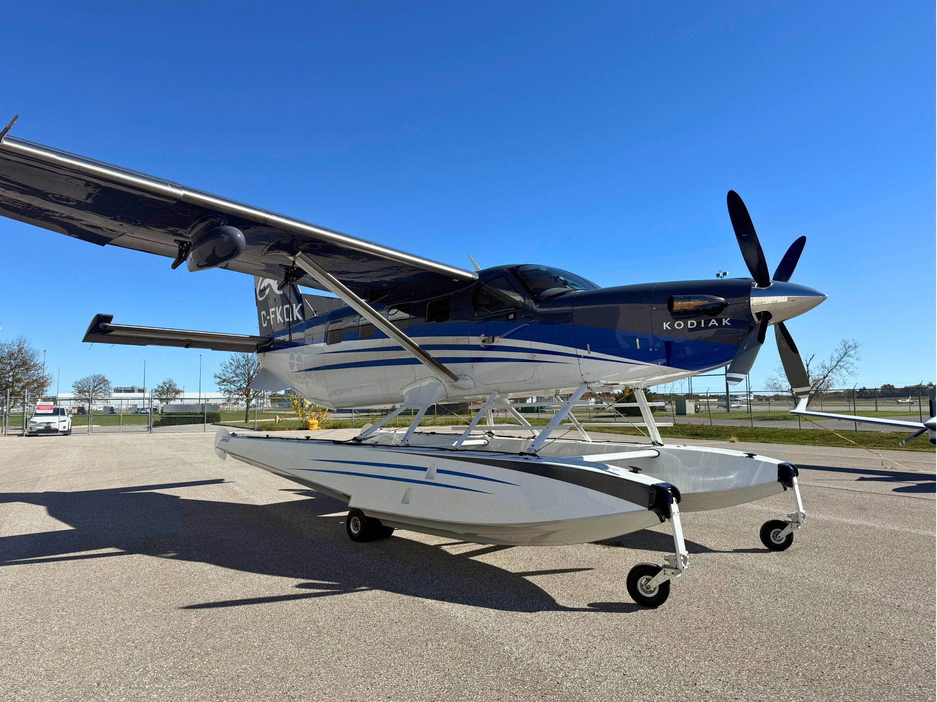 A blue and white Kodiak floatplane on landing gear attached to a wheeled cradle at an airport with clear blue sky above