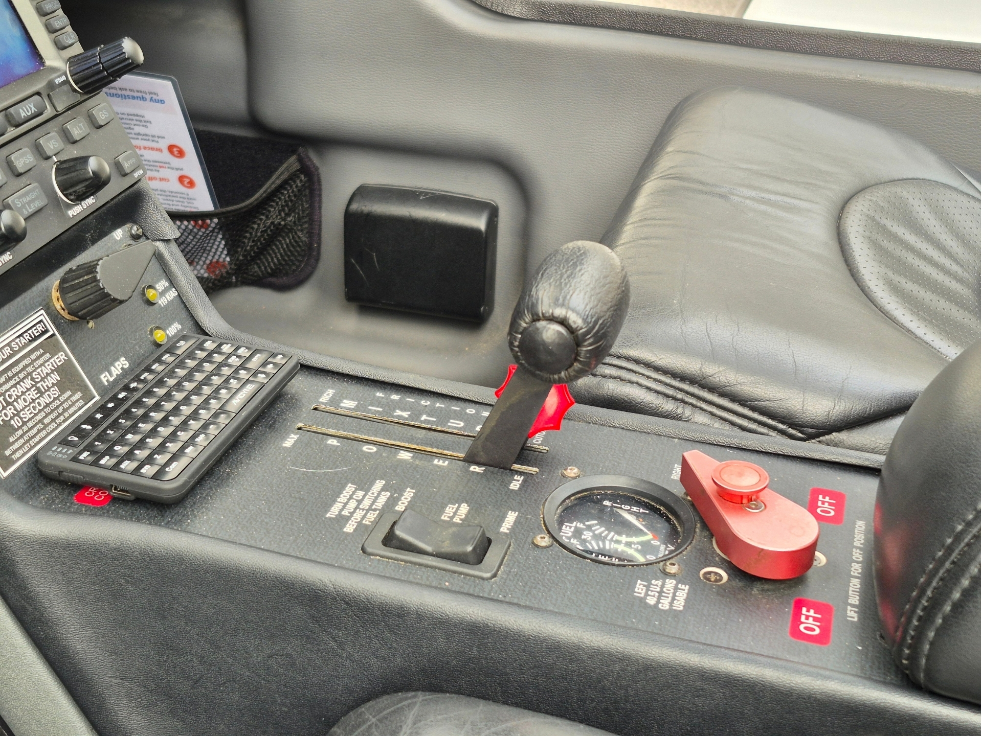 Aircraft cockpit control console with radio throttle lever and red emergency switch next to a round gauge and black leather seat armrest