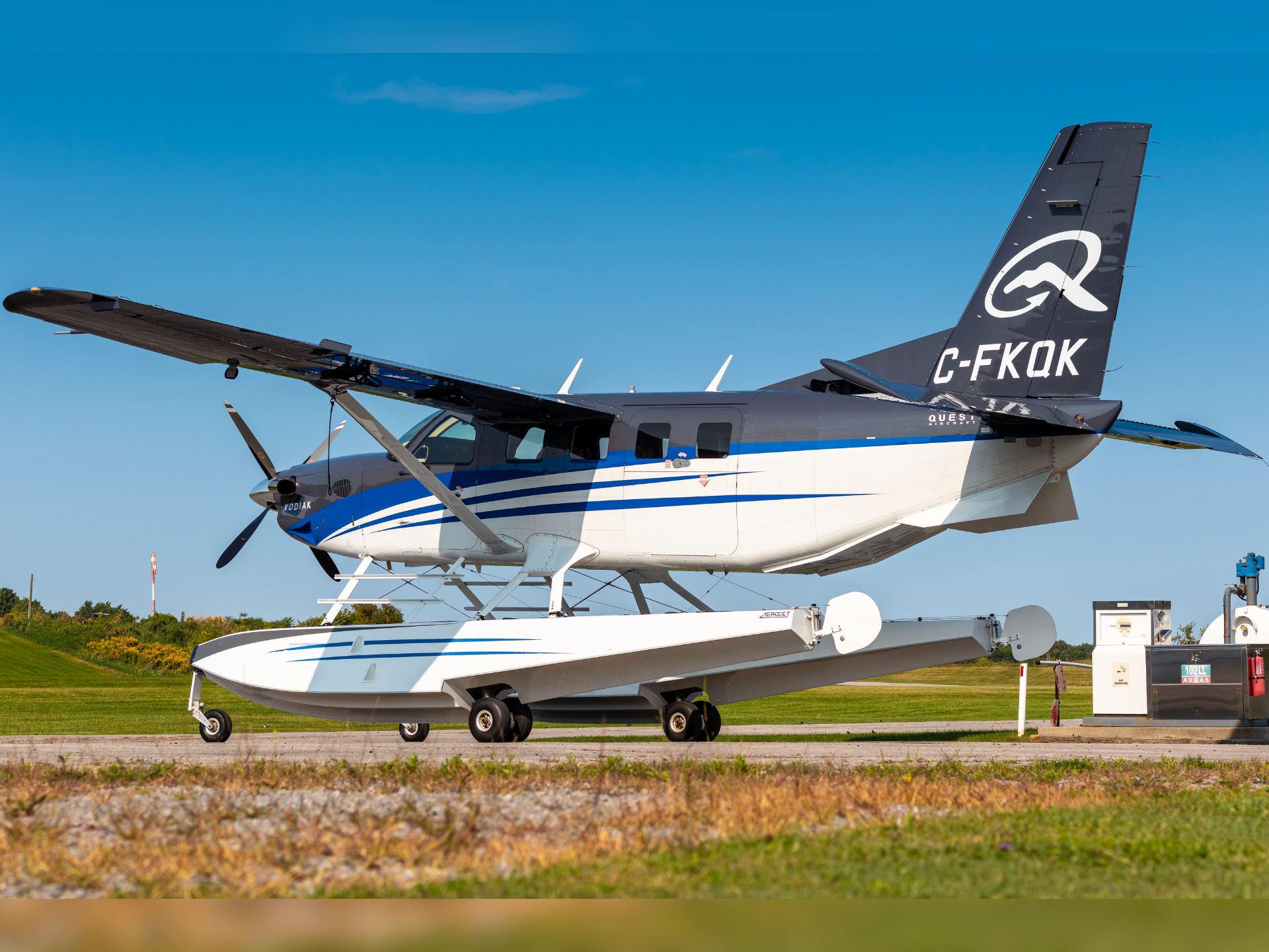 Seaplane on wheels white and blue with a large tail logo parked on a tarmac against a clear blue sky registration C FKQK