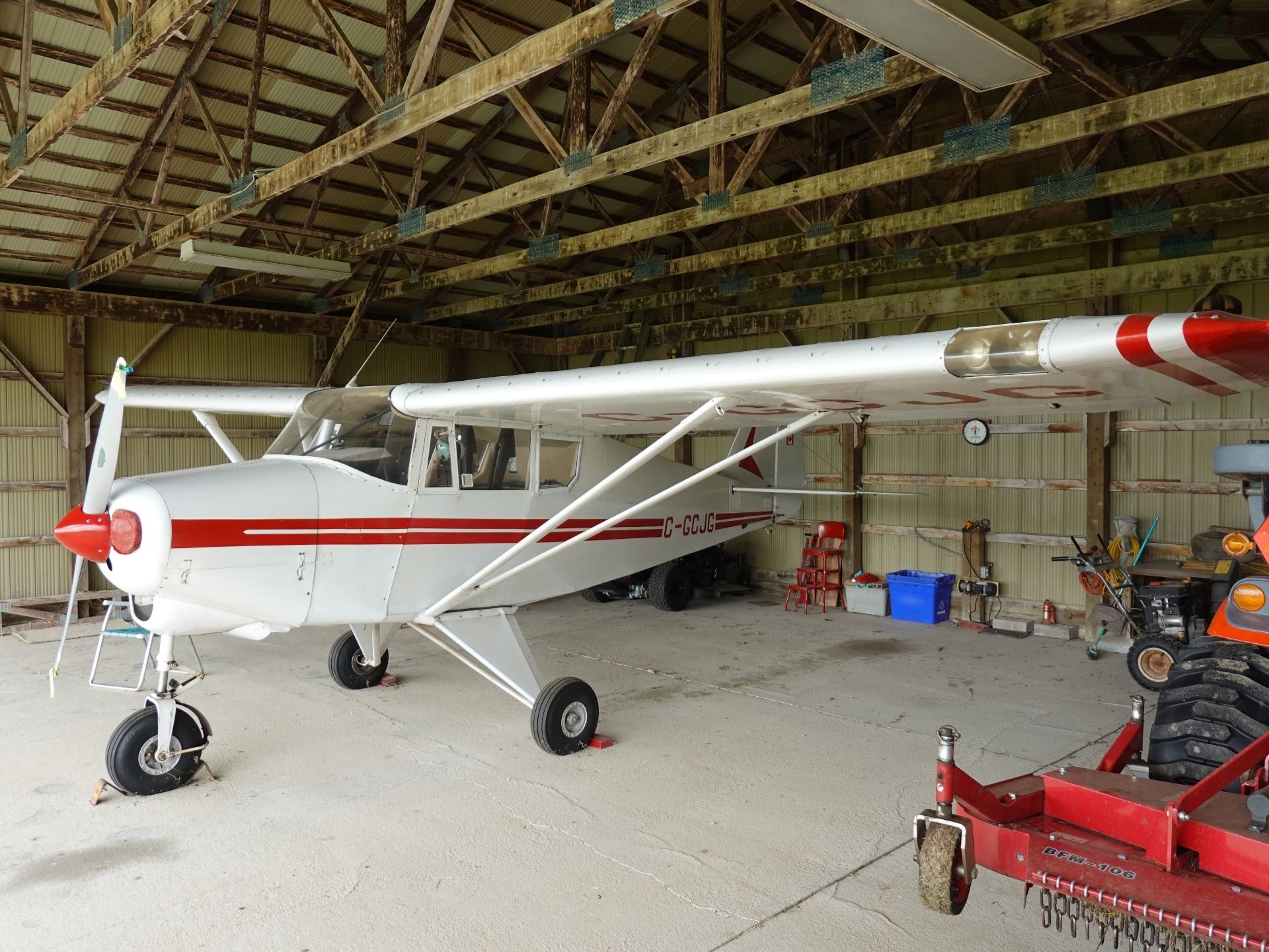 White single-engine airplane with a red nose parked inside a hangar under exposed wooden beams.