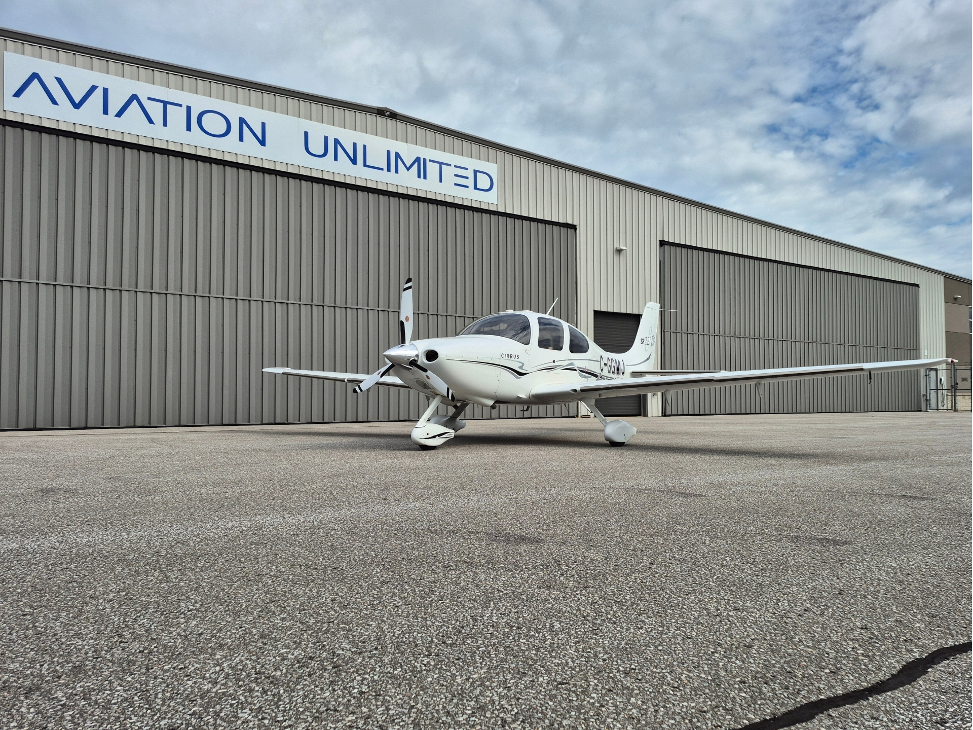 White Cirrus single engine airplane parked on the tarmac in front of a gray hangar with an Aviation Unlimited sign under a cloudy sky