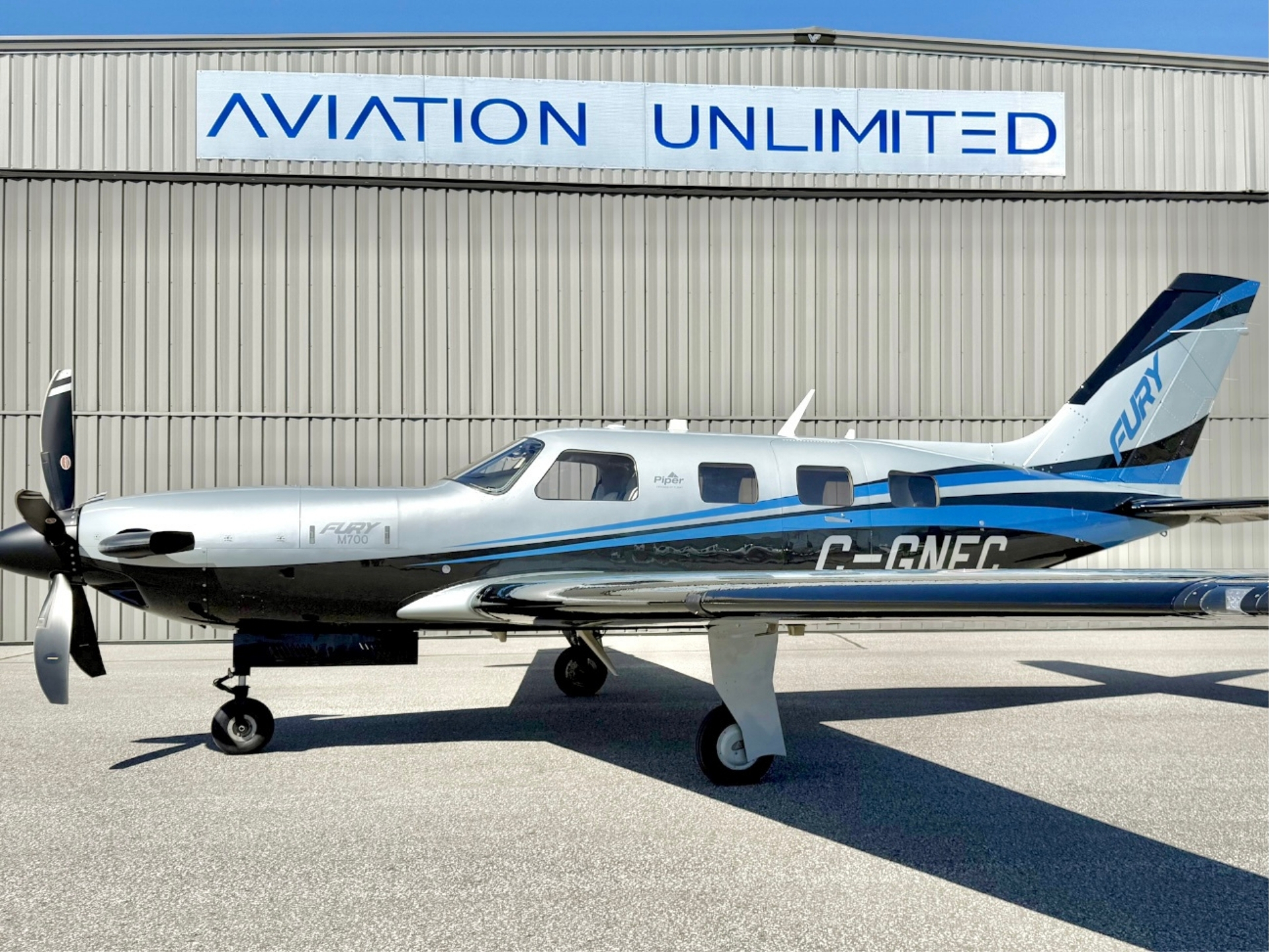 Piper twin engine airplane parked on a tarmac in front of a beige hangar with a blue and white AVIATION UNLIMITED sign above