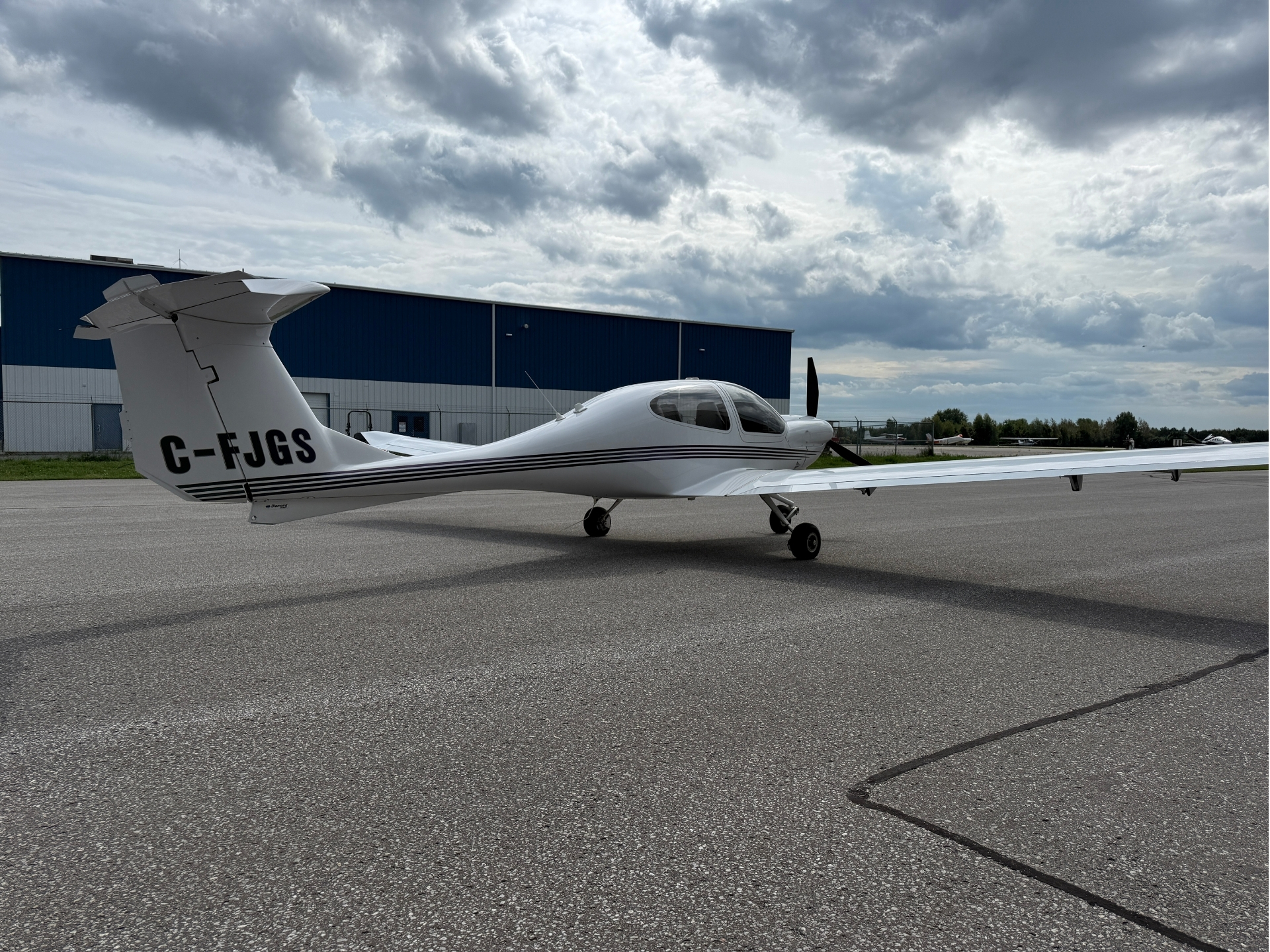 Small white single-engine airplane parked on the tarmac beside a blue hangar, tail number C-FJGS, under a cloudy sky.