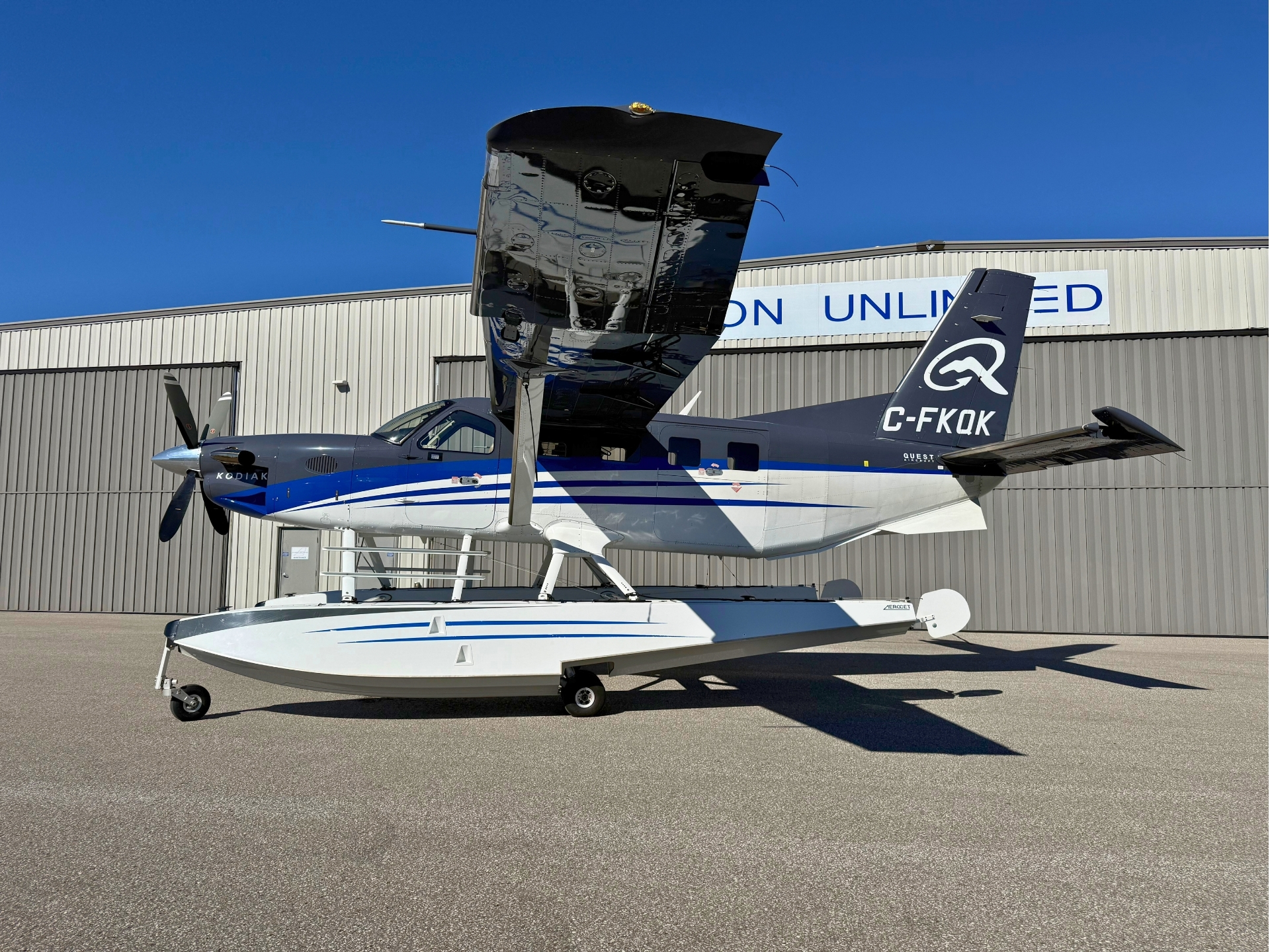 Amphibious airplane on a hull float white and blue livery parked on a tarmac near a hangar with a blue sky the wing is raised above the fuselage