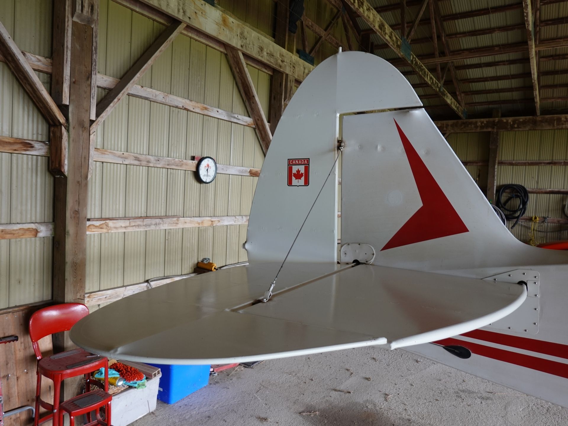 Small aircraft in a wooden hangar, tail fin with red triangle and Canada sticker; cluttered work area with red chairs nearby.