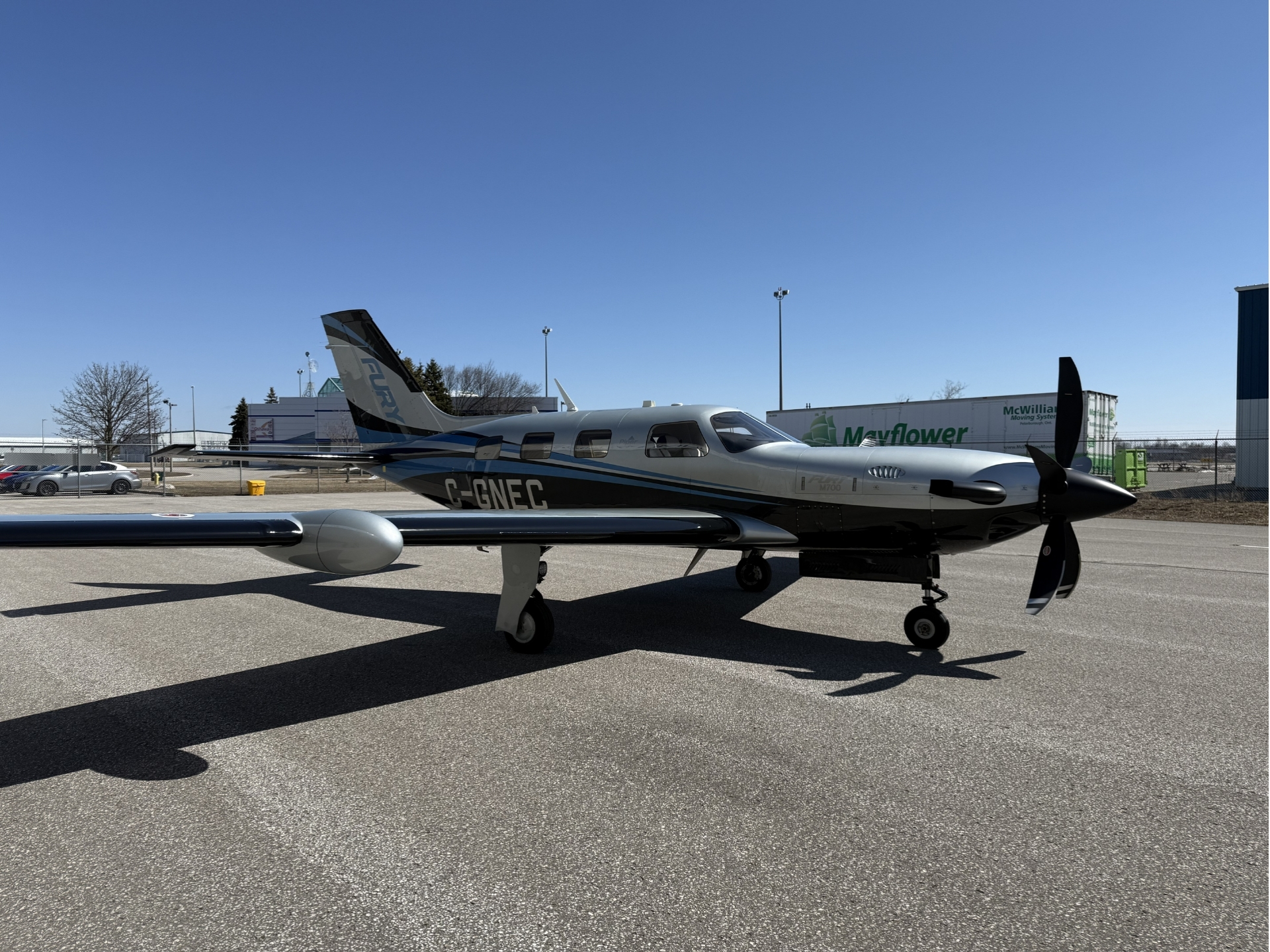 Small twin engine propeller aircraft parked on the tarmac with a clear blue sky tail number C GNEC near hangars in the background