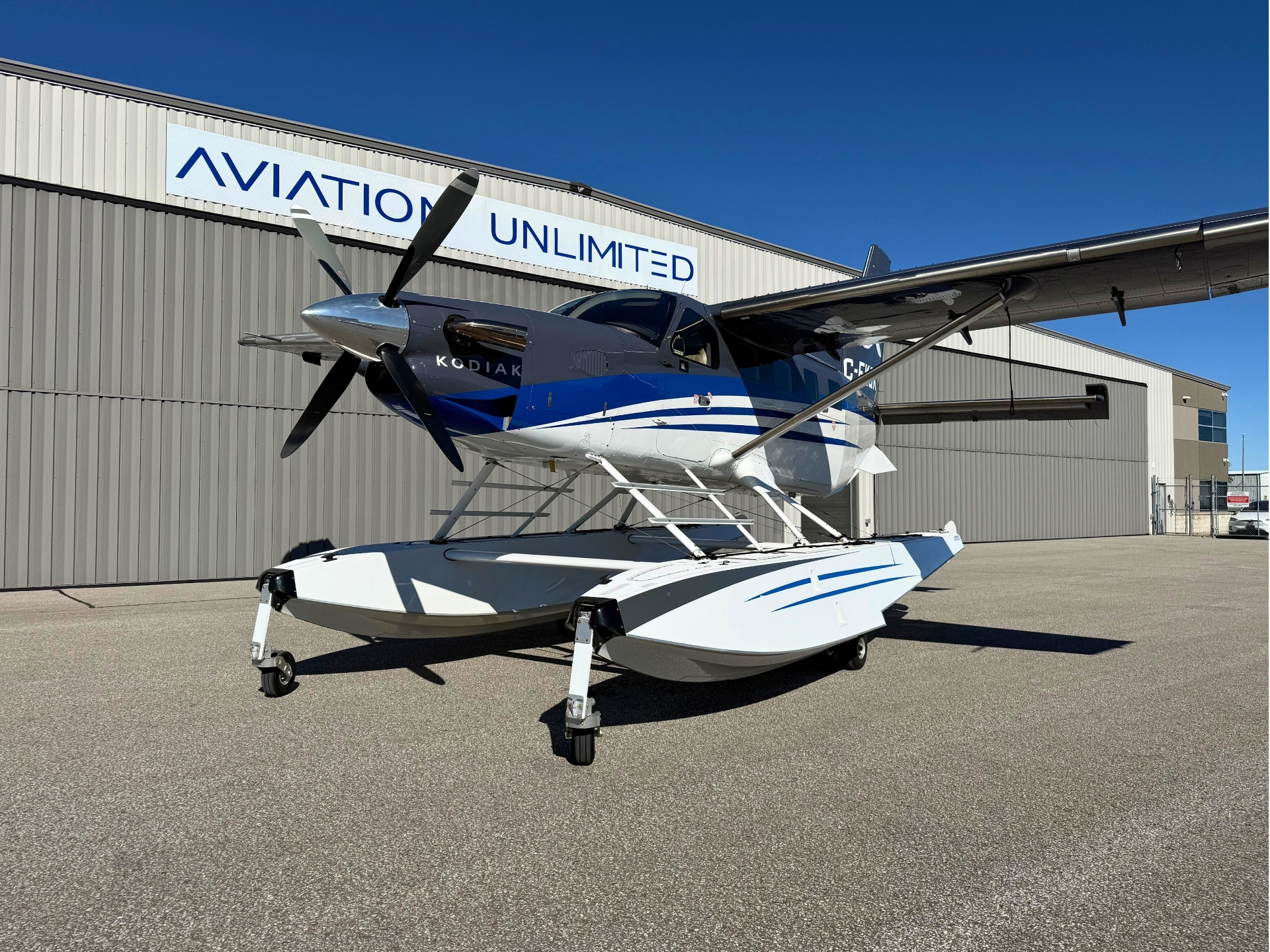 Blue and white Kodiak seaplane on amphibious floats parked in front of a hangar with Aviation Unlimited signage under a bright blue sky
