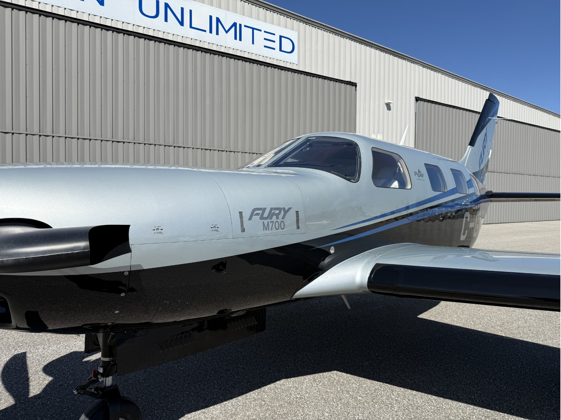 Front left side of a silver M700 aircraft parked on tarmac in front of a gray hangar with a blue UNLIMITED sign showing cockpit windows and propeller