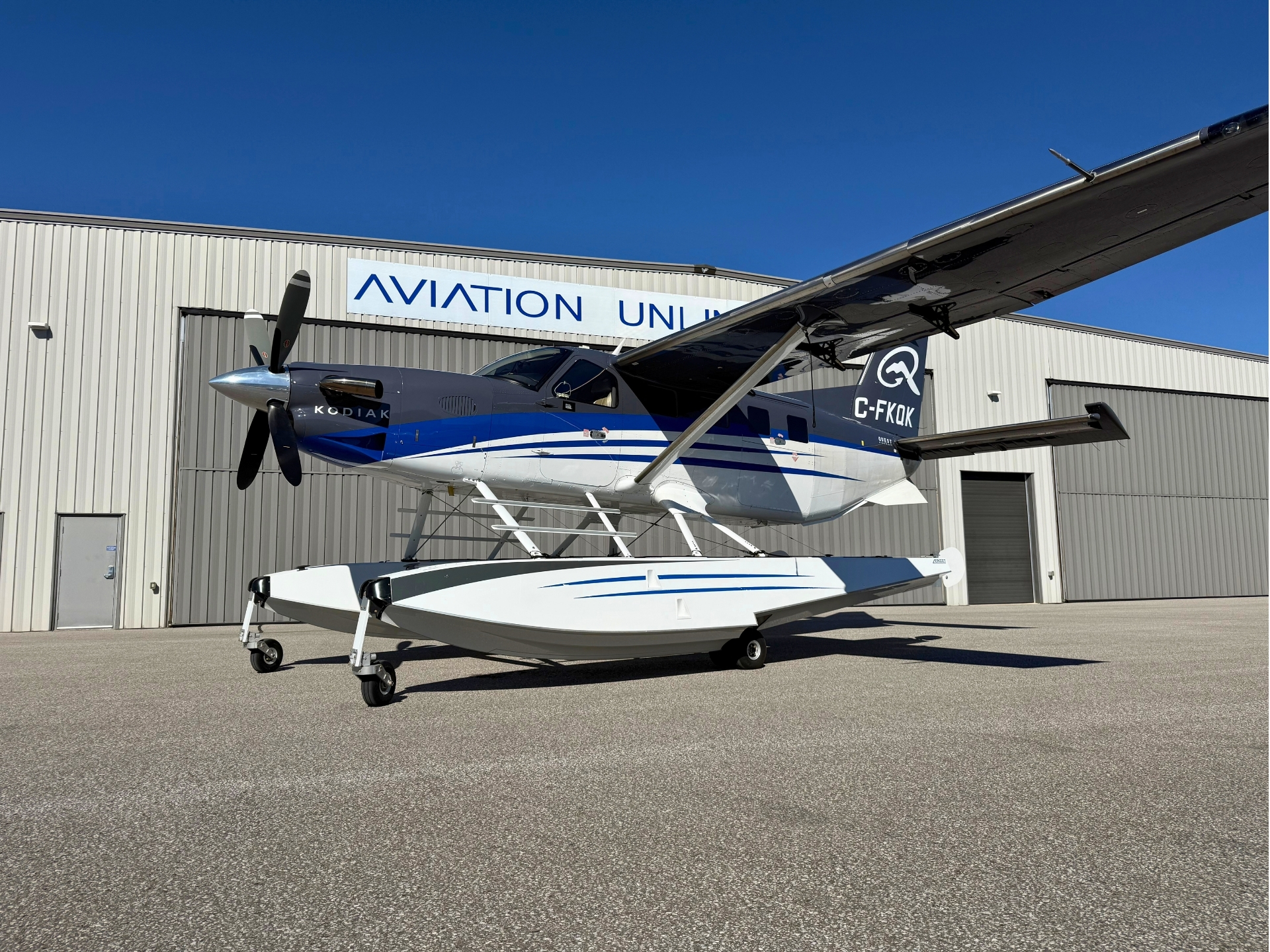 Seaplane on wheels parked on tarmac in front of a corrugated metal hangar with a blue and white paint scheme