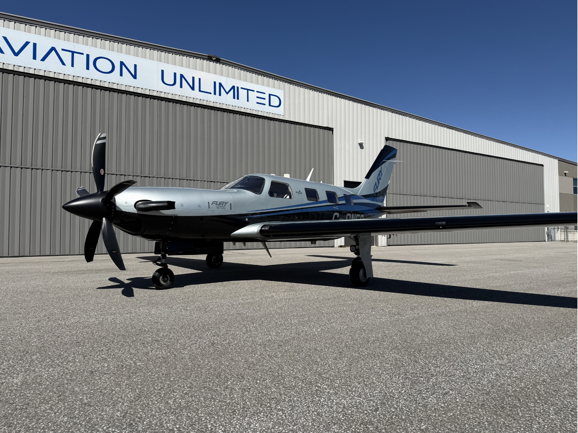 Small gray propeller aircraft with blue accents parked on a tarmac in front of a corrugated metal hangar with a large AVIATION UNLIMITED sign against a clear blue sky