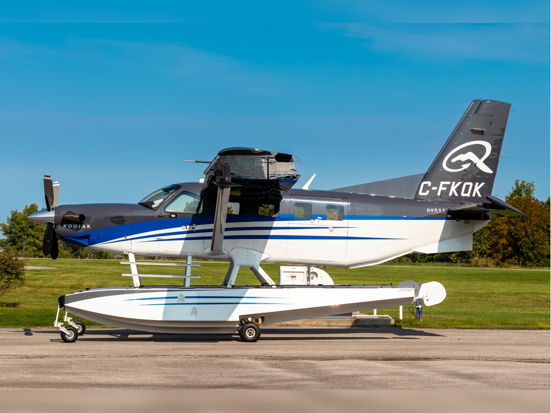 Side view of a gray and white Quest Kodiak amphibious airplane with floats tail number C FKOK parked on a runway with a clear blue sky