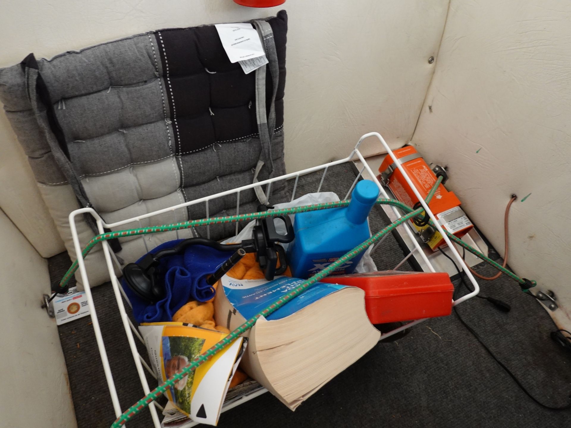 A cluttered storage area inside a small space: cushions, magazines, a blue container, and an orange tool box mixed with hoses and a white metal rack on the floor.
