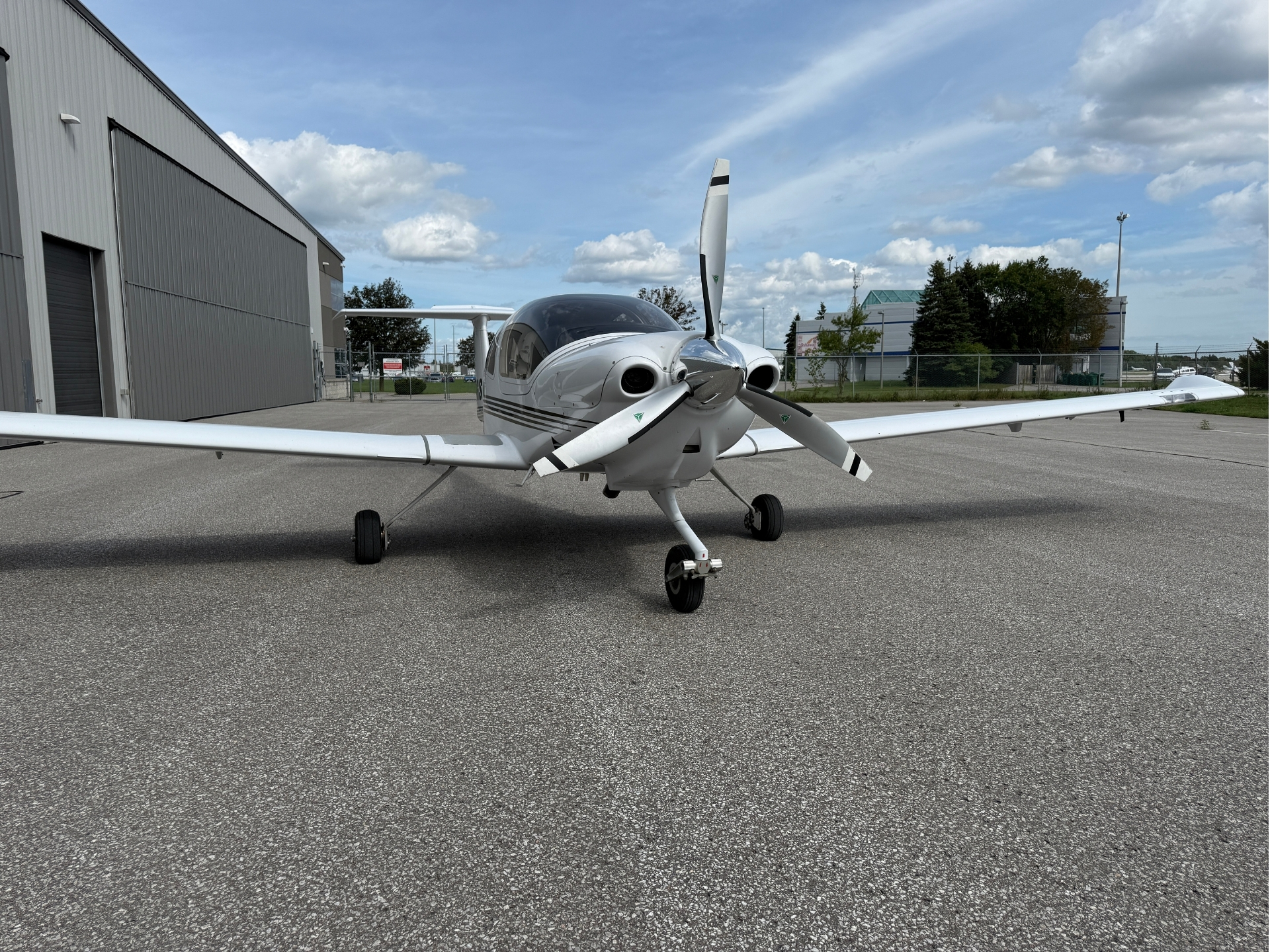 Front view of a silver twin-engine propeller airplane parked on a paved tarmac beside a hangar under a blue sky with clouds.