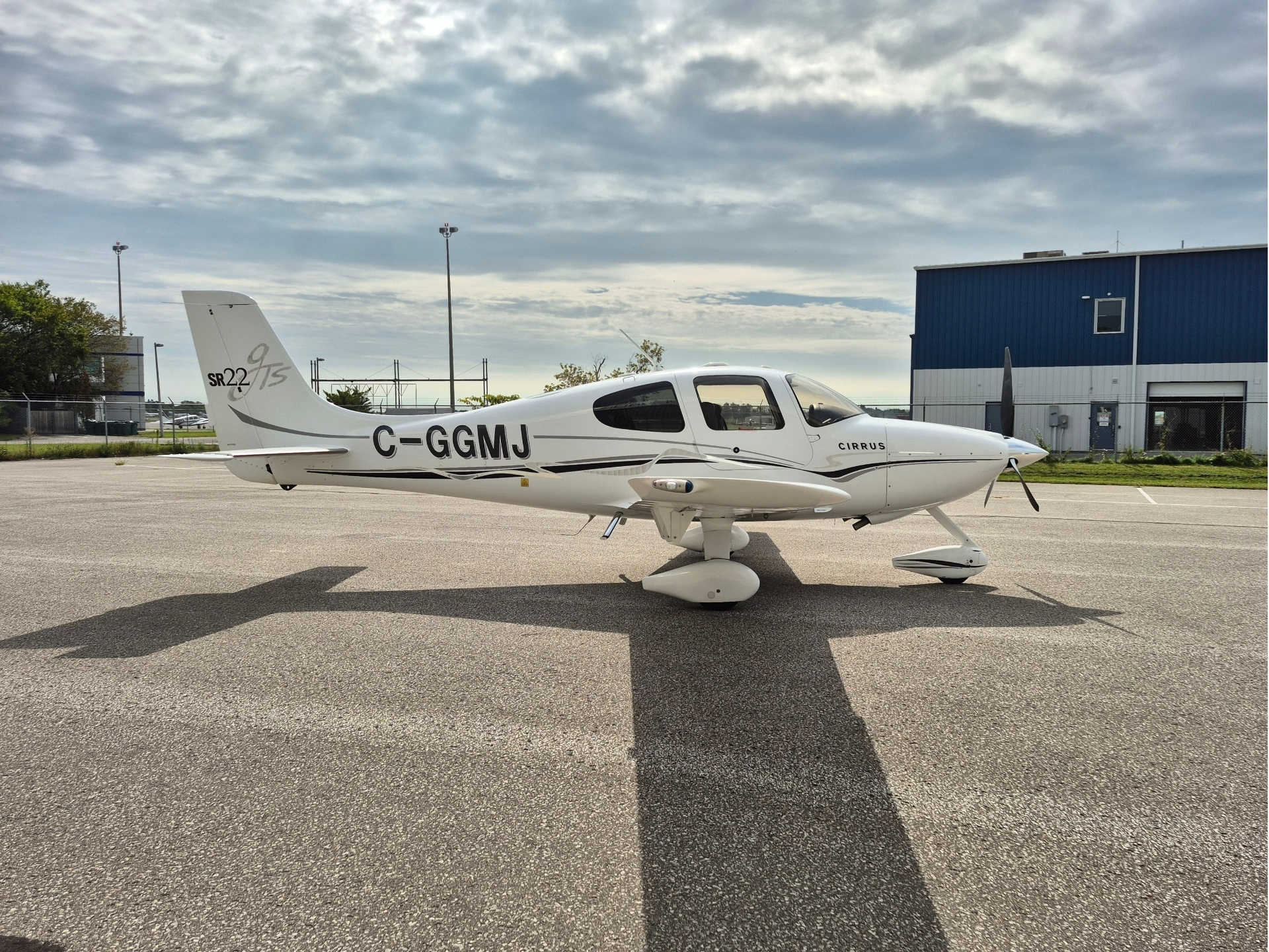 White Cirrus SR22 airplane with registration C GGMJ parked on the airport tarmac blue hangar in the background under a cloudy sky