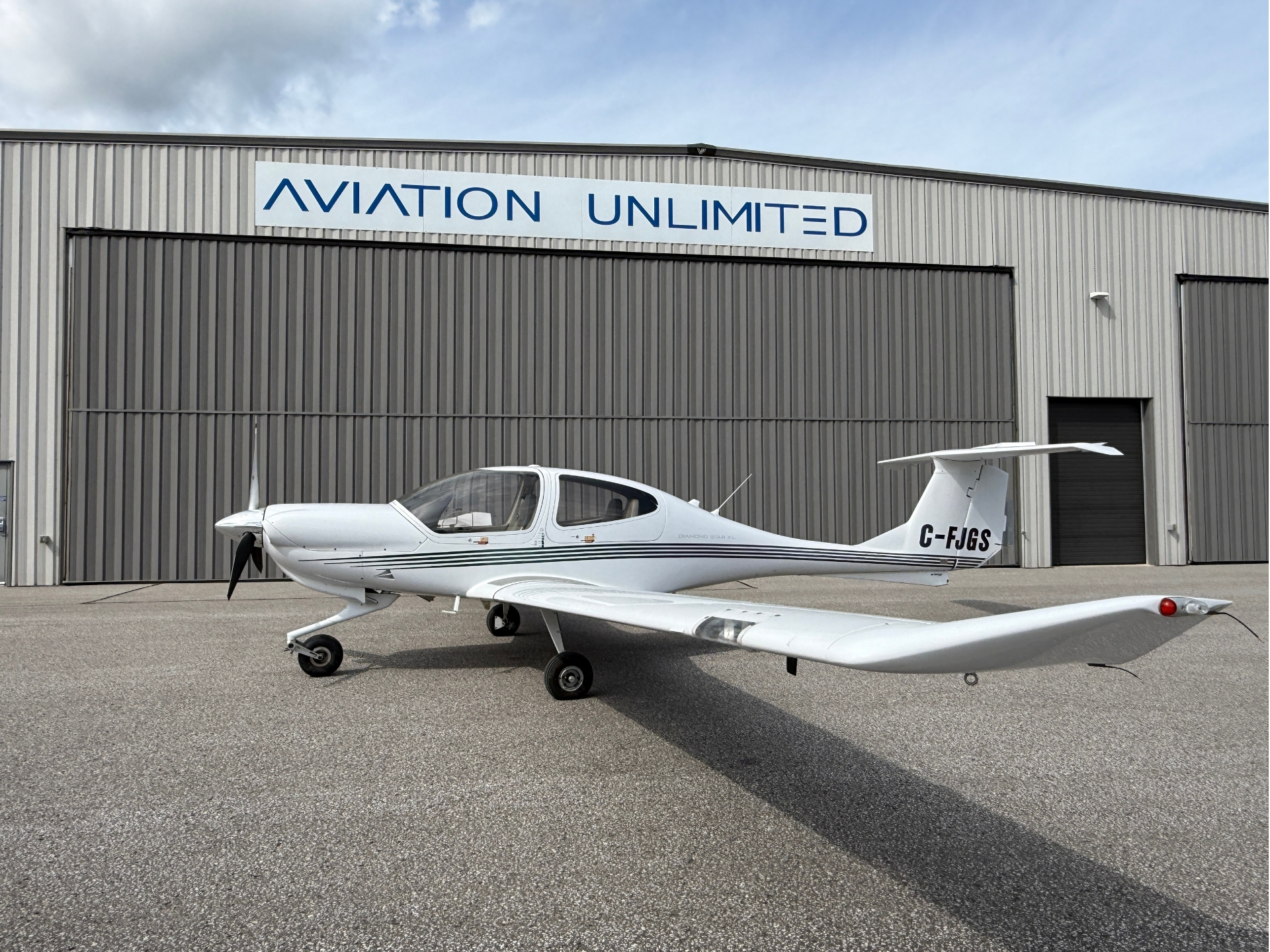 White single‑engine propeller airplane parked on a tarmac in front of a gray corrugated hangar with an ‘AVIATION UNLIMITED’ sign above the door, tail number C-FJGS visible.