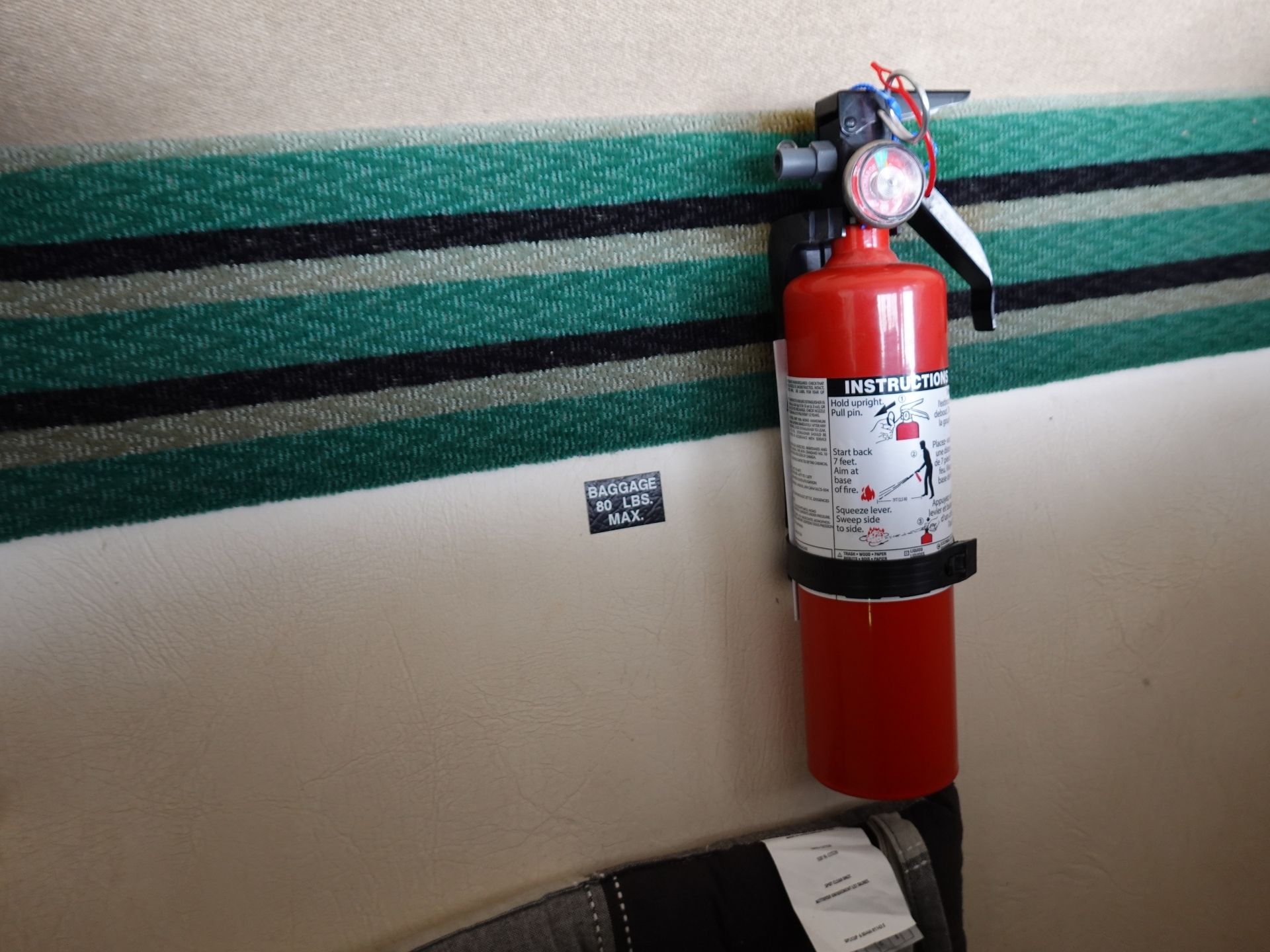 Red fire extinguisher mounted on a beige wall with a green striped rug behind and instruction label visible.