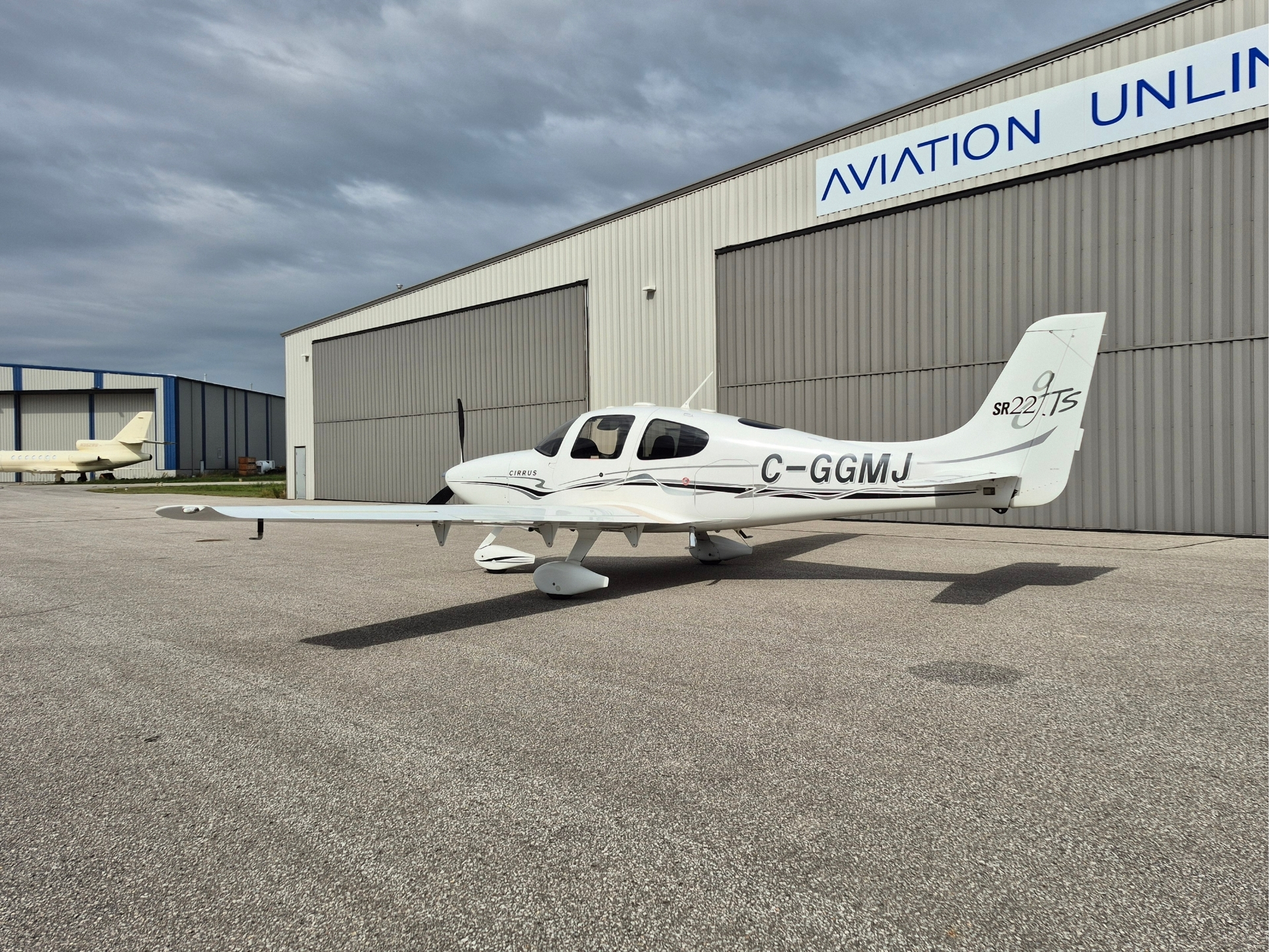White Cirrus SR22 aircraft parked on the tarmac before a large aviation hangar under a cloudy sky