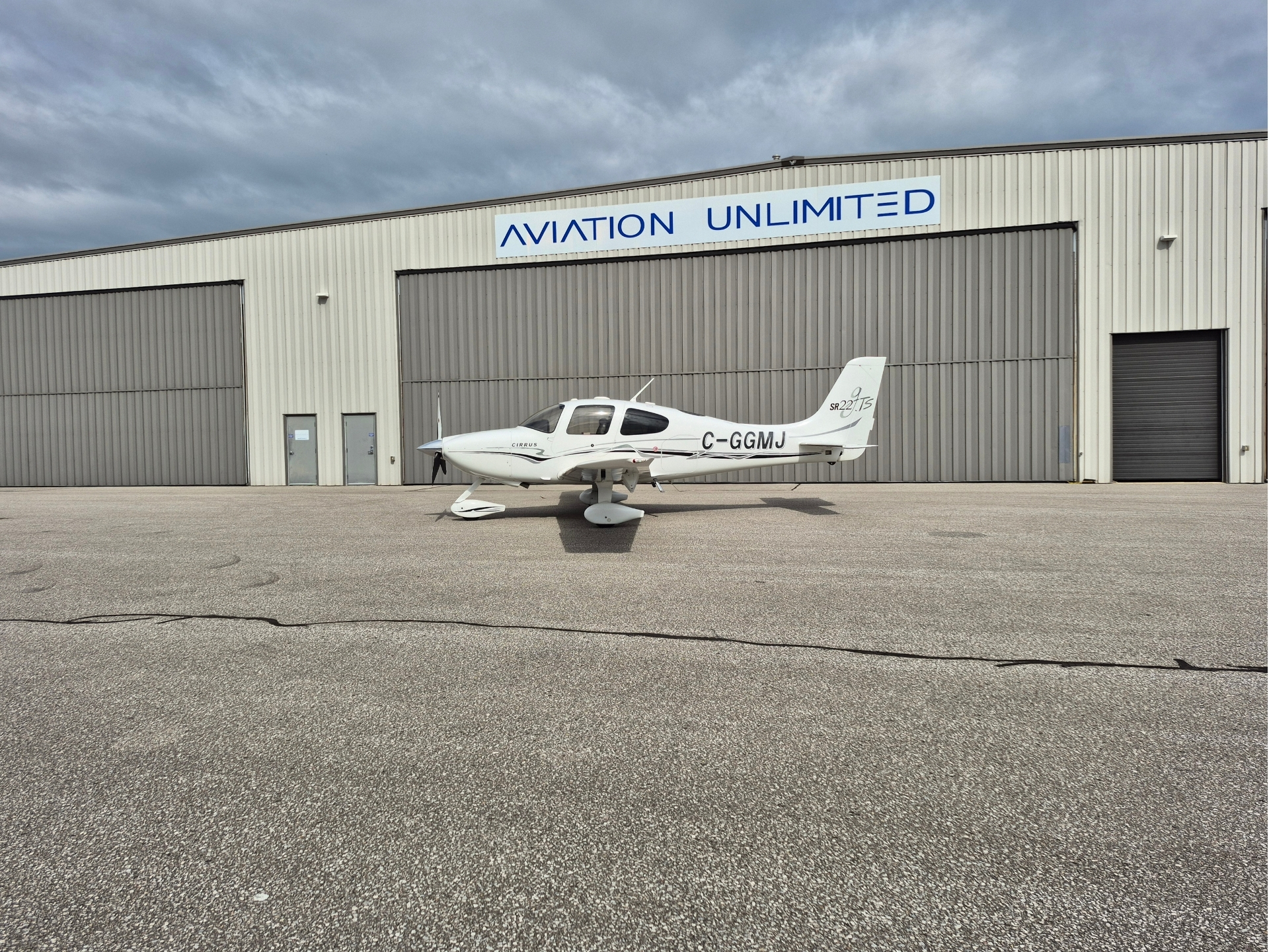 White Cirrus SR22 airplane parked on the tarmac in front of a large hangar with a blue Aviation Unlimited sign above the doors under a cloudy sky
