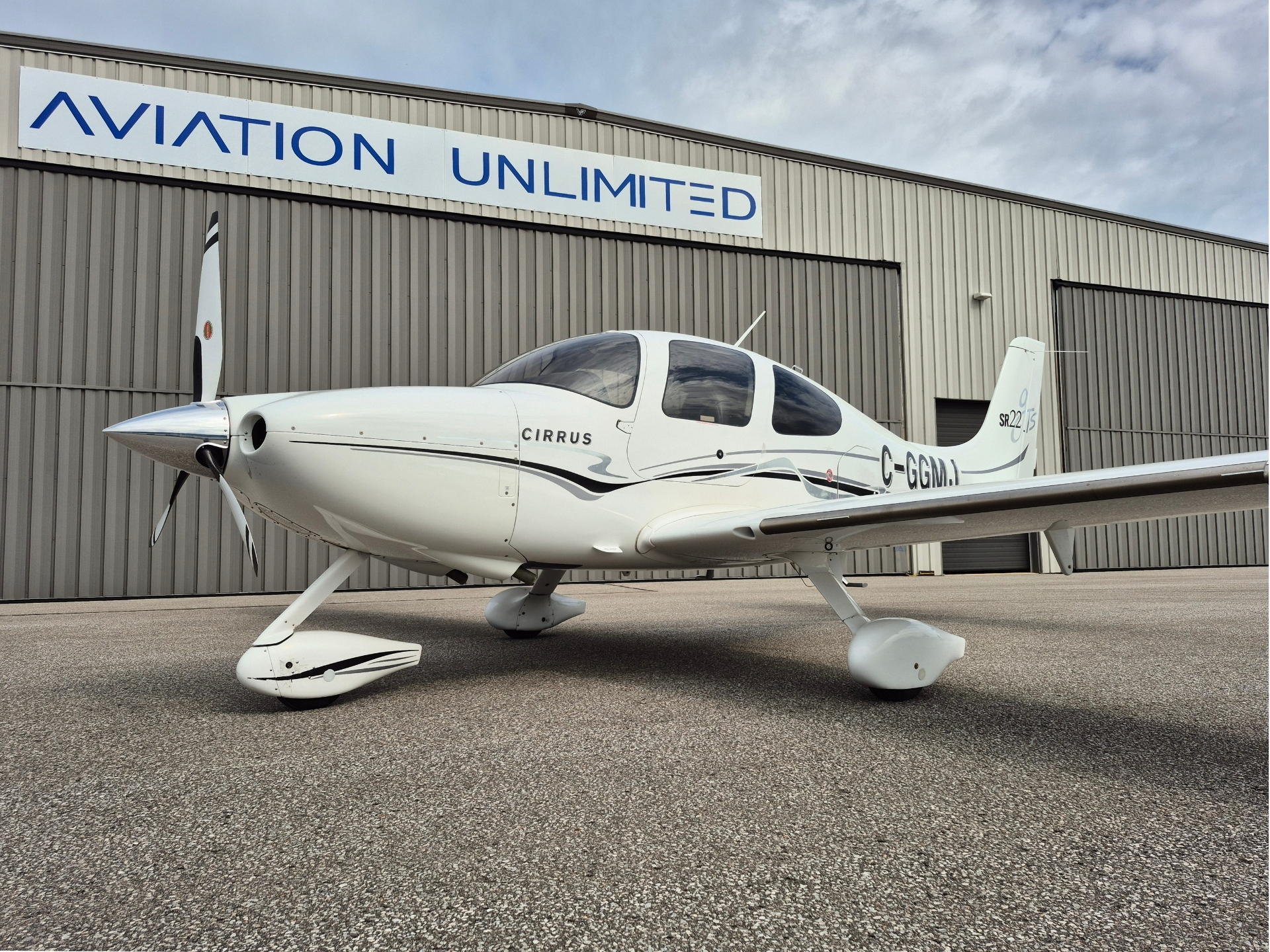 White Cirrus single engine airplane parked on the tarmac in front of a gray hangar with a blue Aviation Unlimited sign